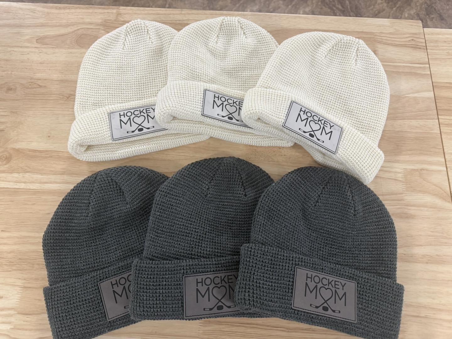 Six knit beanies, three in cream color and three in dark gray, arranged in two rows on a wooden surface. Each beanie has a patch that says ‘Hockey MOM’ with a heart, crossed hockey sticks, and a small ball.
