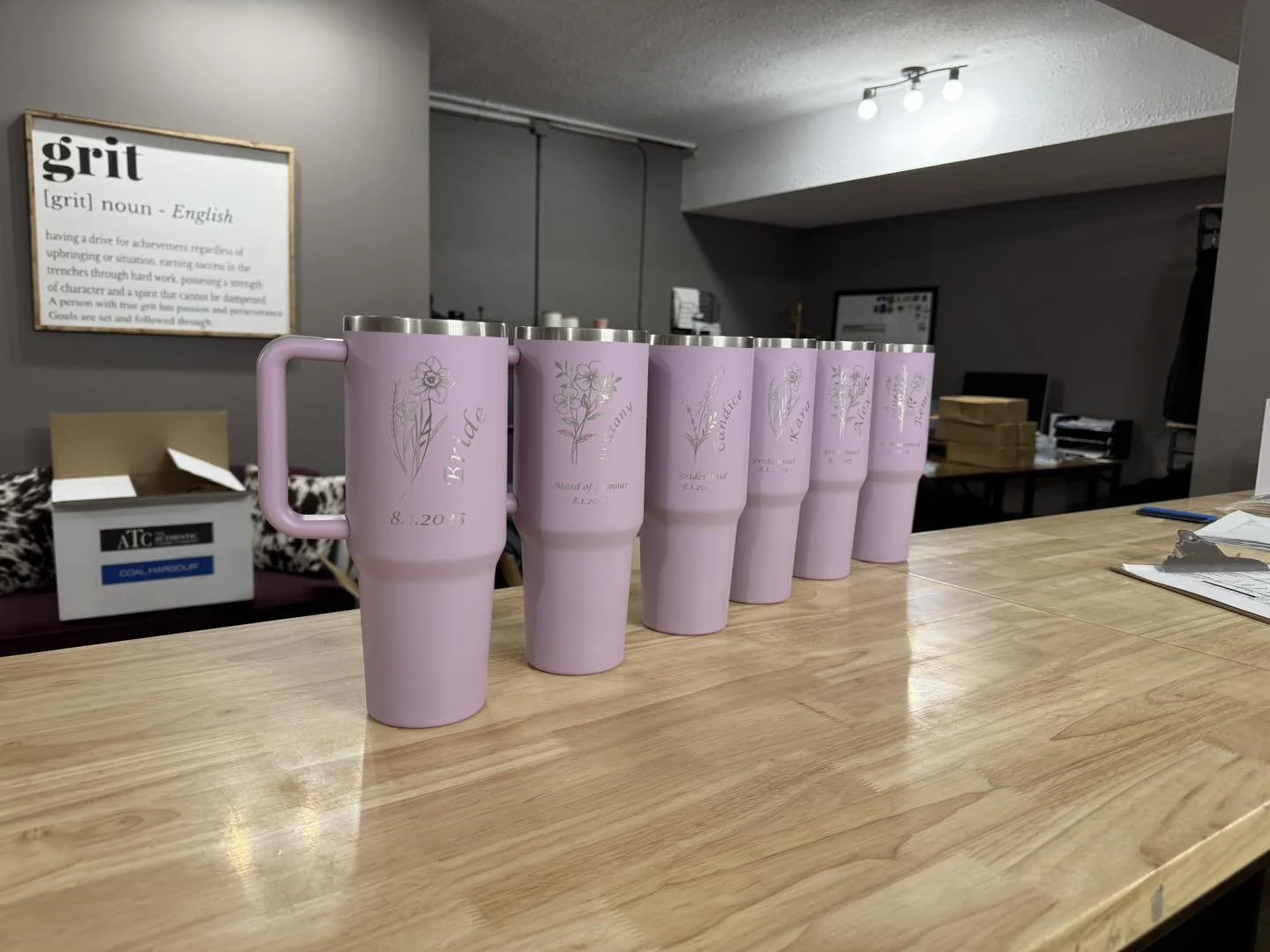 Six pink personalized tumblers with floral designs and engraved names on a wooden table in an indoor setting.