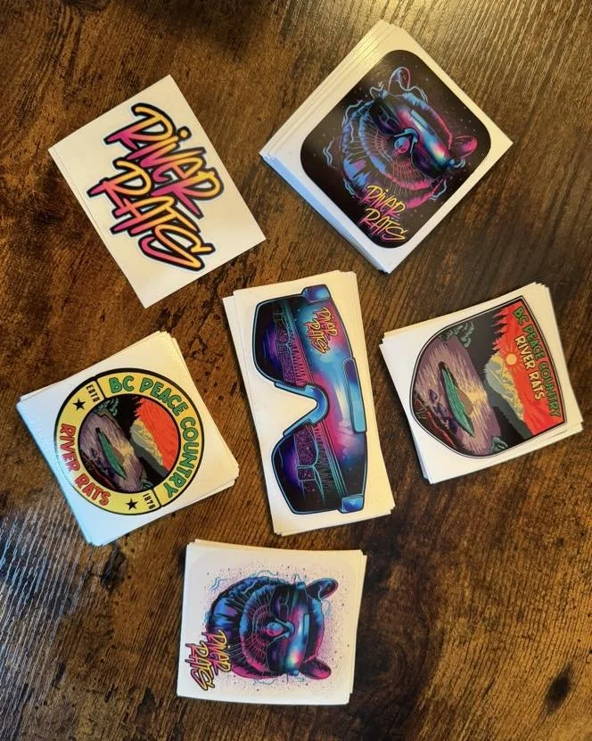 Collection of sticker sheets and individual stickers with 1980s neon style graphics, including sunglasses, a fish, and a landscape scene, on a wooden table.