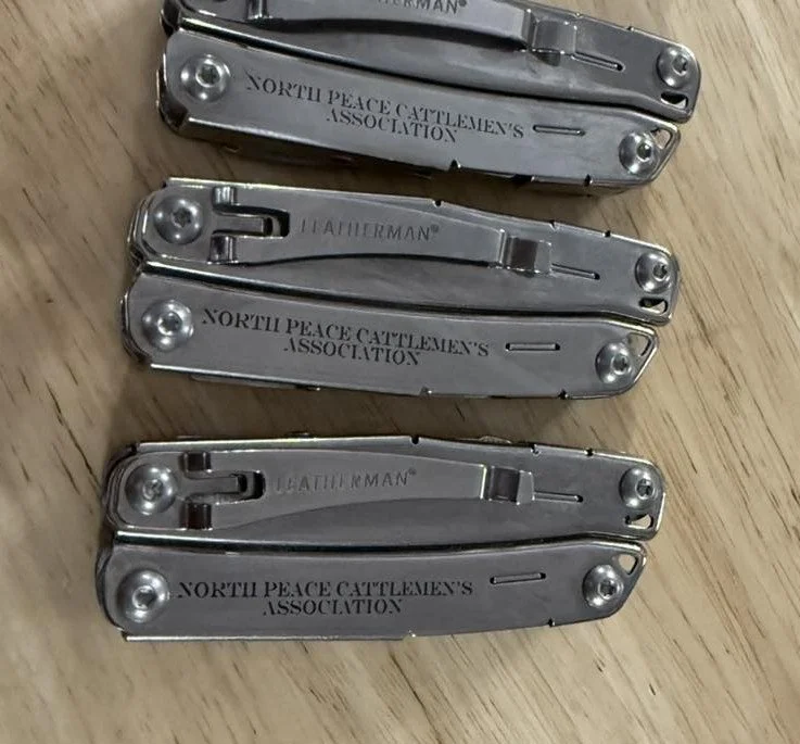 Four metal multi-tool knives on a wooden surface, with the top item inscribed with 'North Peace Cattlemen's Association' and the second with 'Leatherman'.