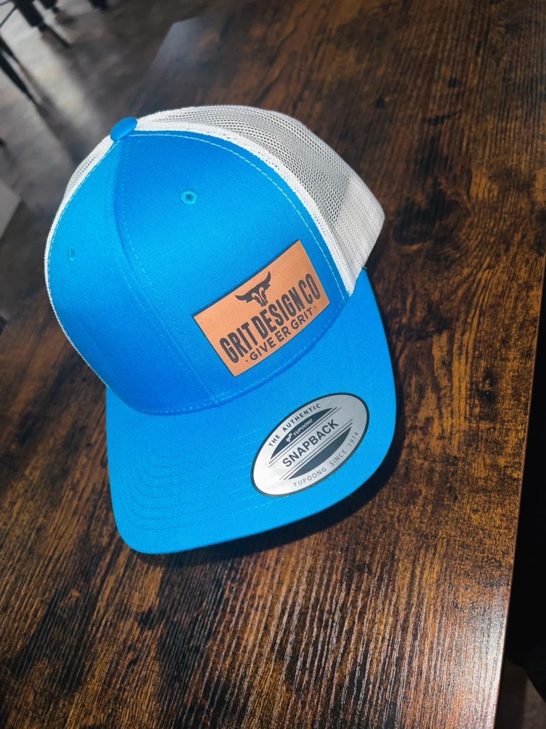 A blue and white snapback cap with a leather patch reading 'GRIT DESIGN CO GIVE ER GRIT' and a sticker that says 'The Authentic Snapback Yupoong Since 1972' on the bill, resting on a wooden surface.
