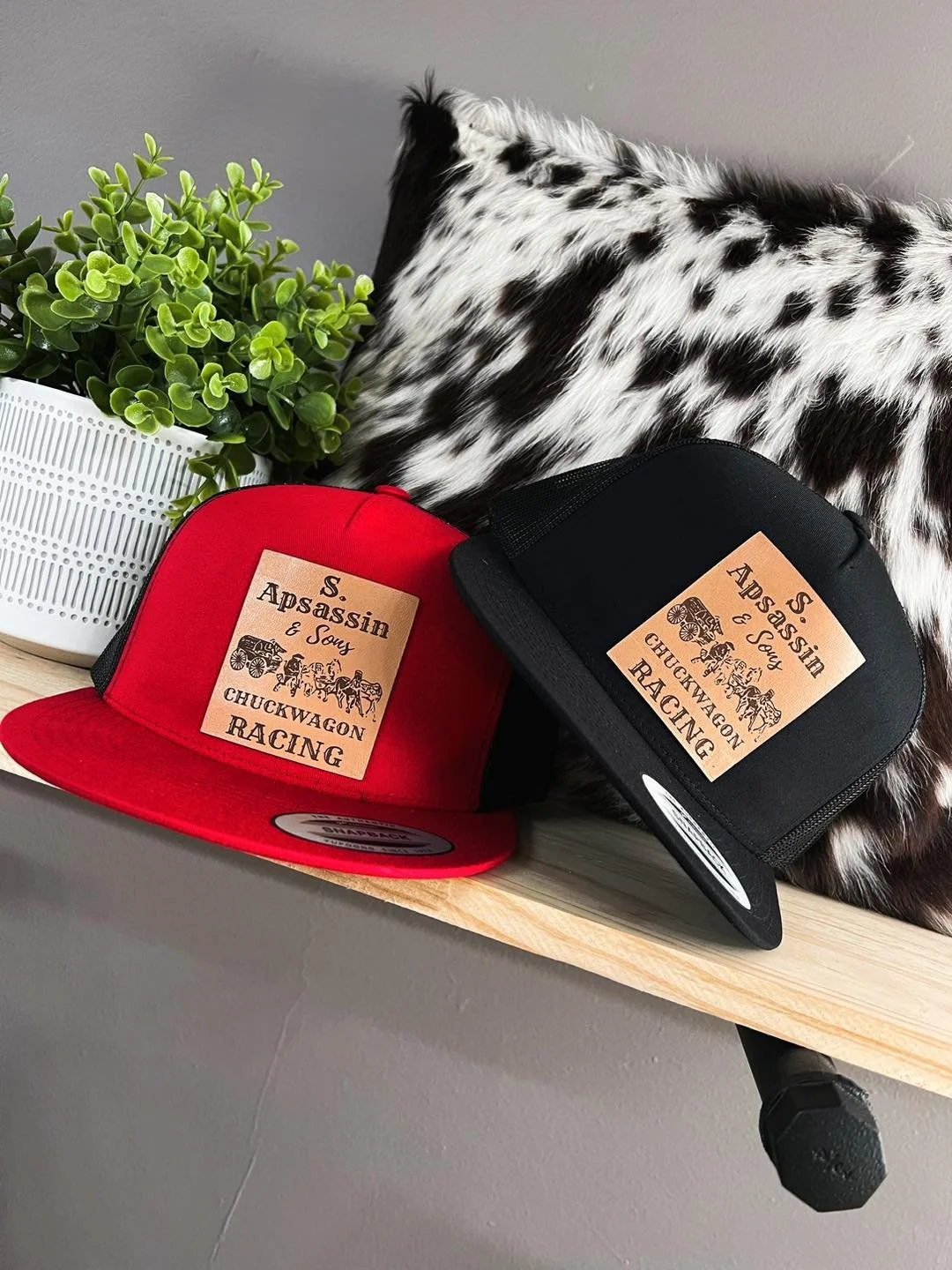 Red and black snapback hats with a leather patch reading 'S. Upsassin & Souy, Chuckwagon Racing,' placed on a wooden surface next to a green potted plant and a black and white fur pillow.