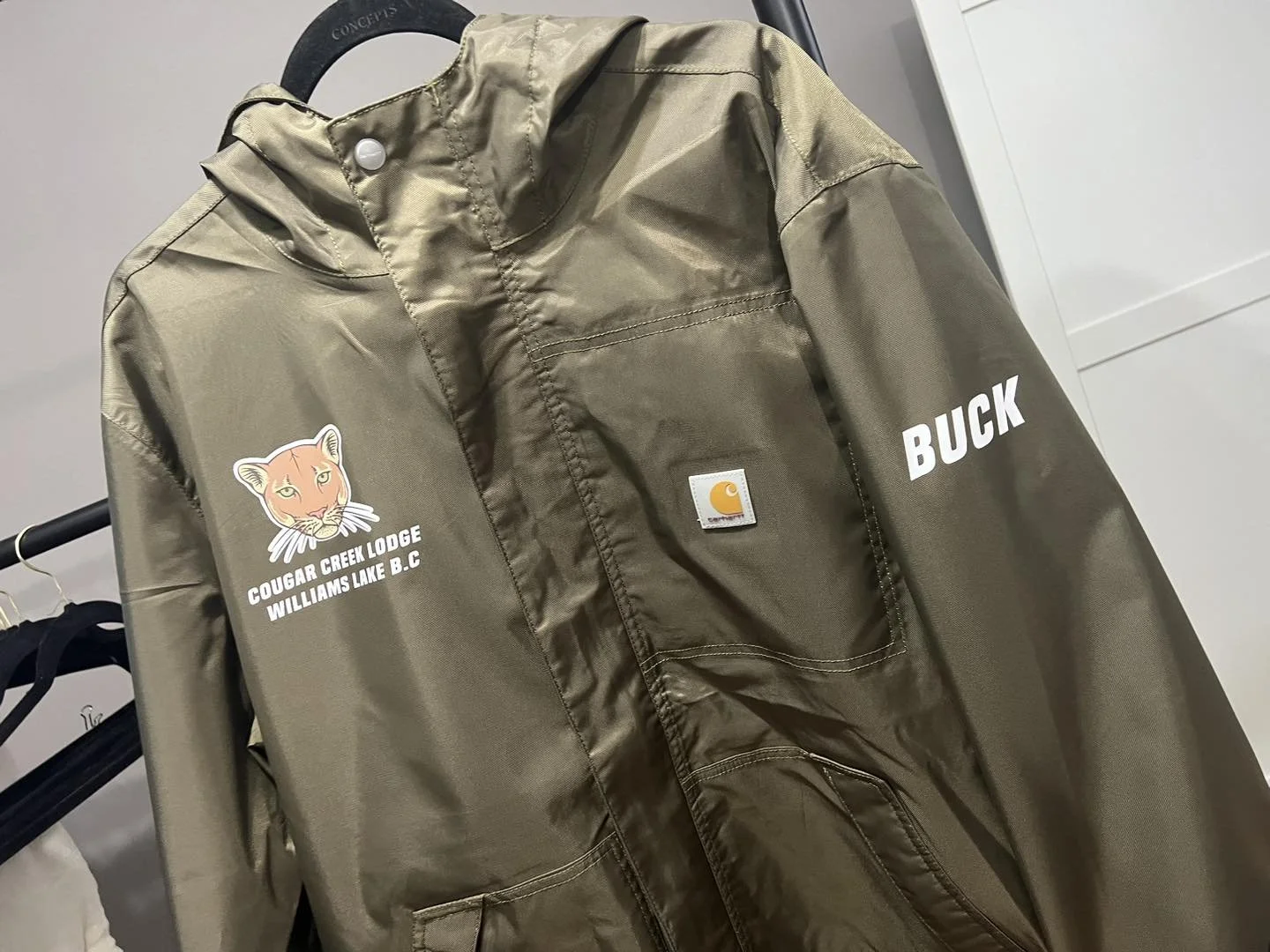A khaki Carhartt jacket hanging on a black hanger. The jacket has a Cougar Creek Lodge logo with a cougar's face and the text "COUGAR CREEK LODGE WILLIAMS LAKE B.C." on the left chest. The right sleeve has the name "BUCK" in white text.