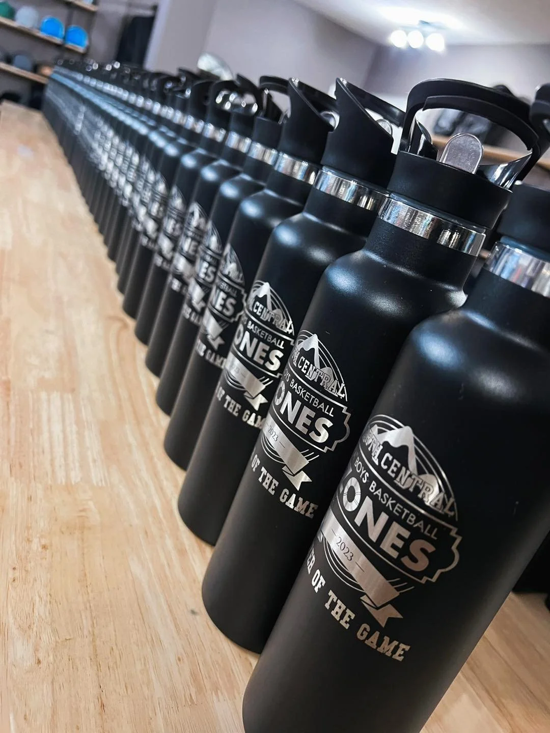A row of black water bottles with handles and silver caps on a wooden surface, each bottle displaying a logo for a basketball game event.