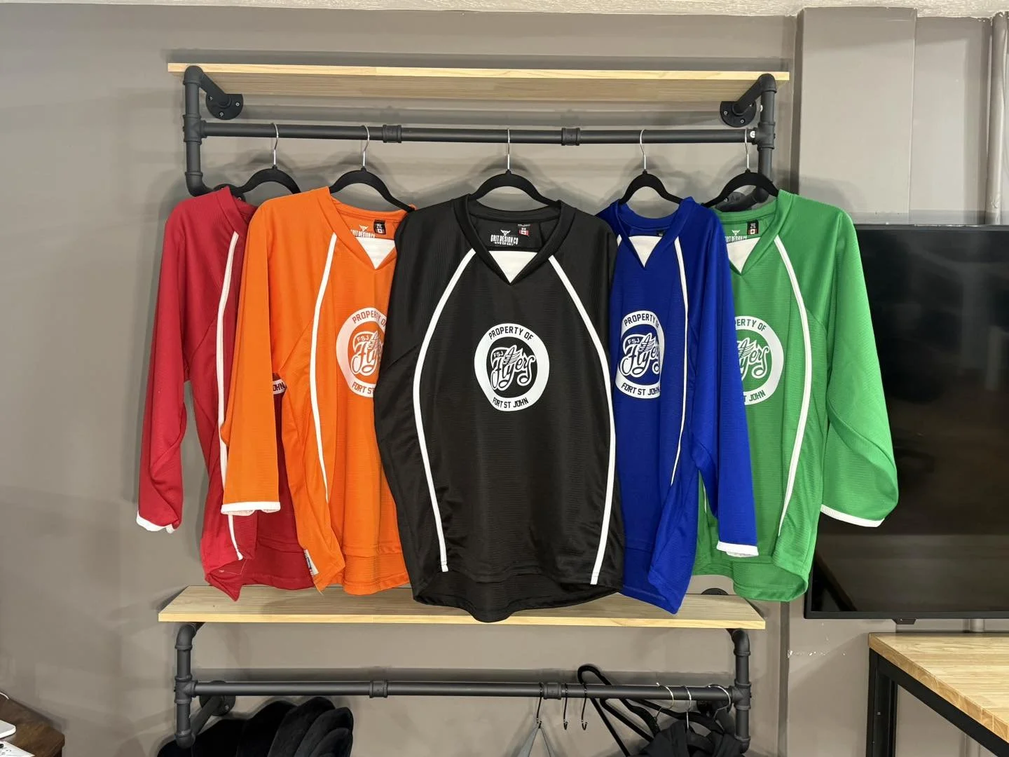 Six colorful sports jerseys hanging on a black metal and wood rack, arranged in red, orange, black, blue, and green colors from left to right.