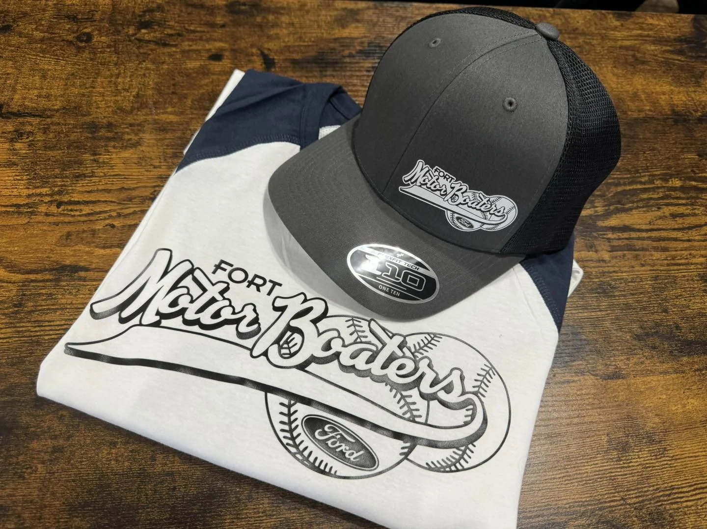 Black and gray Ford Motorcraft baseball cap resting on a folded white Motorcraft T-shirt with a baseball graphic, placed on a wooden surface.