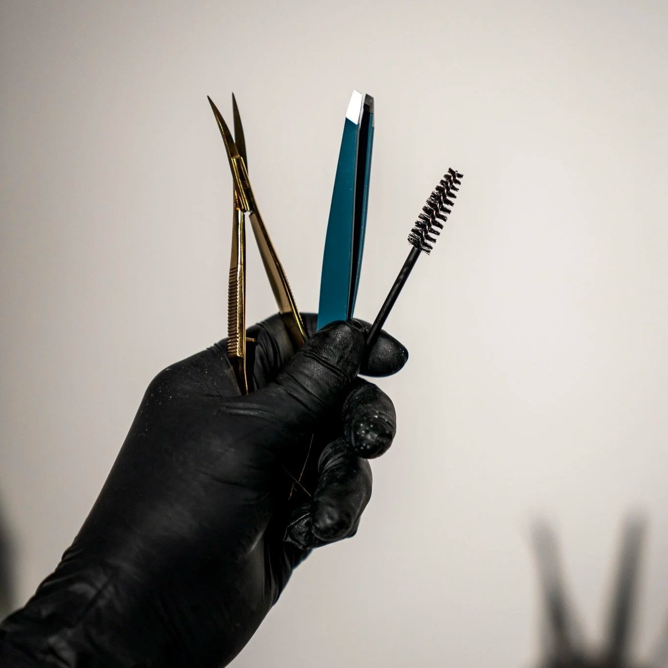 A gloved hand holding a pair of cuticle scissors, a nail clipper, a nail file, and a mascara wand.