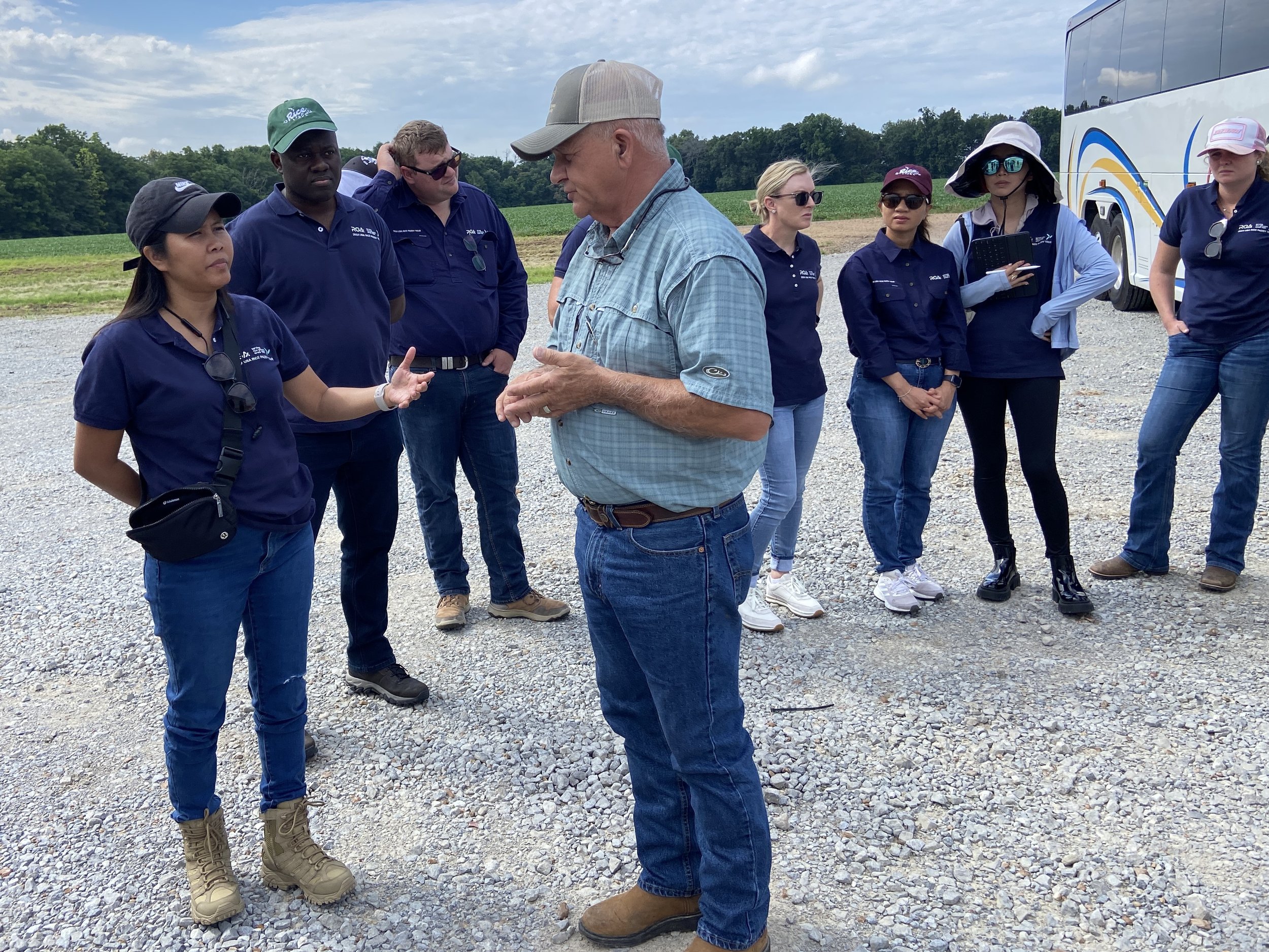 A group of people, mostly women, are gathered outdoors on a gravel area, listening to an older man in a light blue shirt and baseball cap speaking. Some are wearing navy blue shirts, and there's a bus parked nearby with trees and fields in the background.