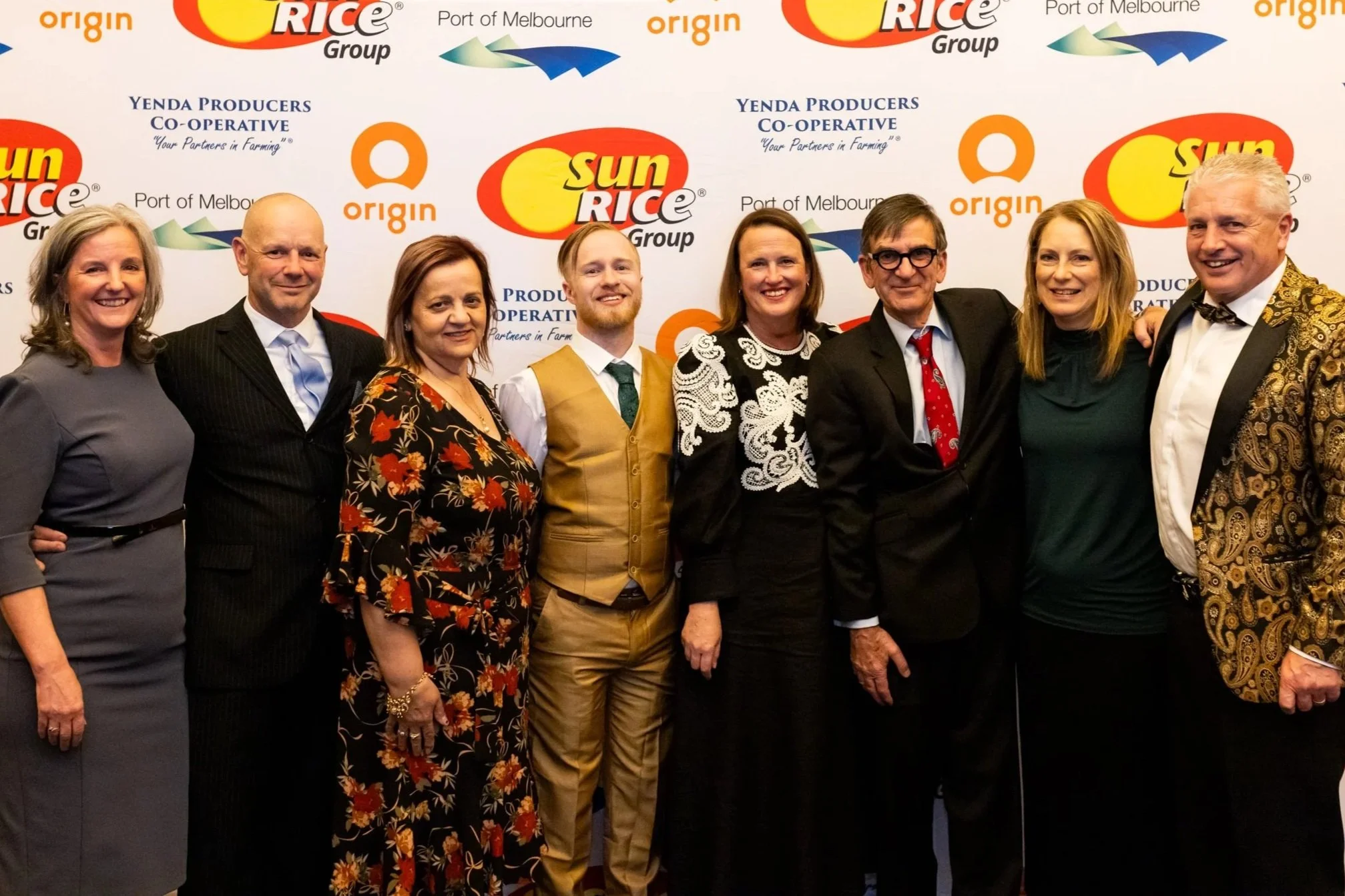 A group of eight people standing and smiling in front of a backdrop with logos and text related to SunRice and other organizations.