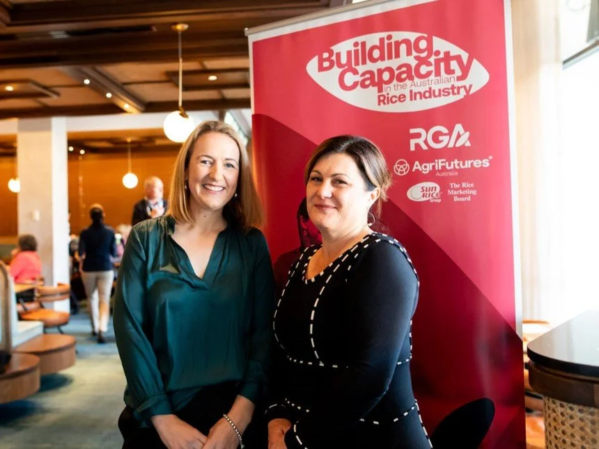 Two women standing in front of a red banner at an indoor event, smiling for the camera.