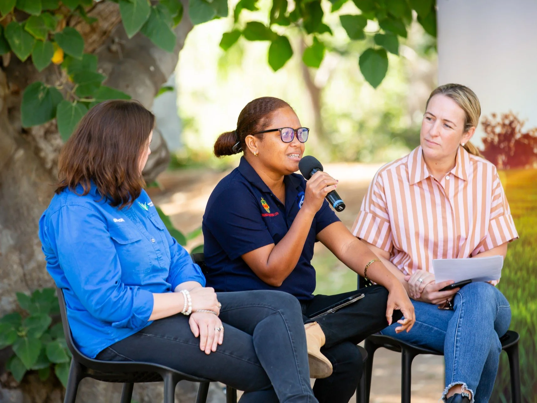 Three women sitting outdoors, engaged in a discussion with one woman speaking into a microphone. The background includes large green leaves and a tree trunk.