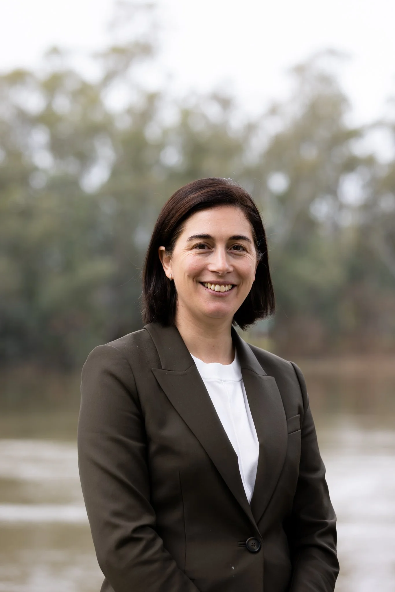 A woman with dark hair outdoors, smiling, wearing a checkered blazer and a dark shirt, surrounded by trees and greenery.