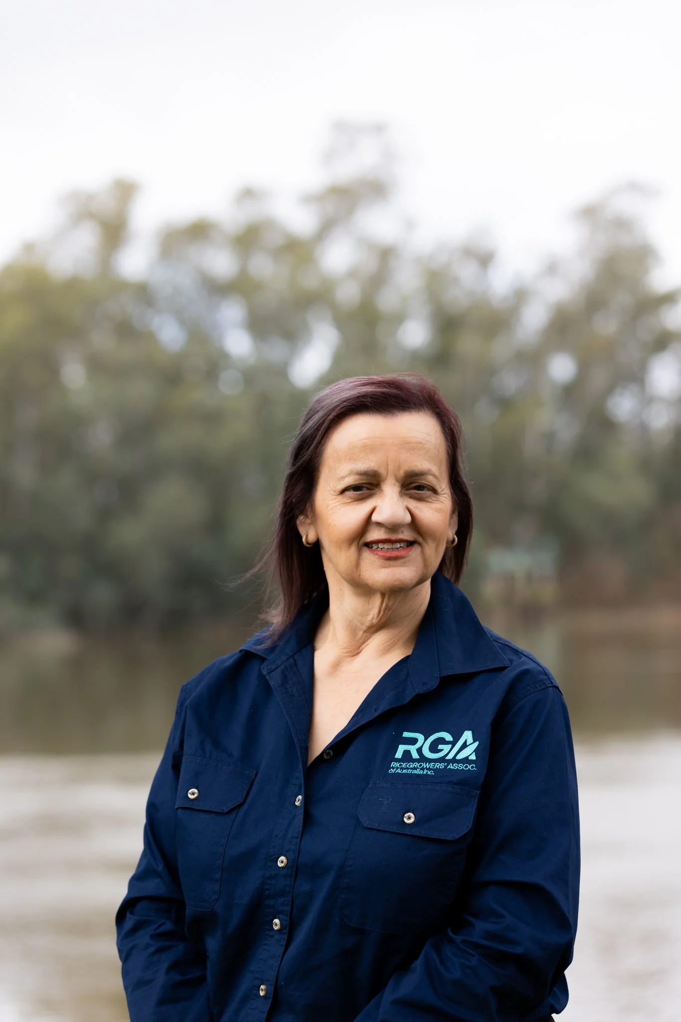 A woman standing outdoors next to a tree with long green leaves, dressed in black, smiling at the camera.