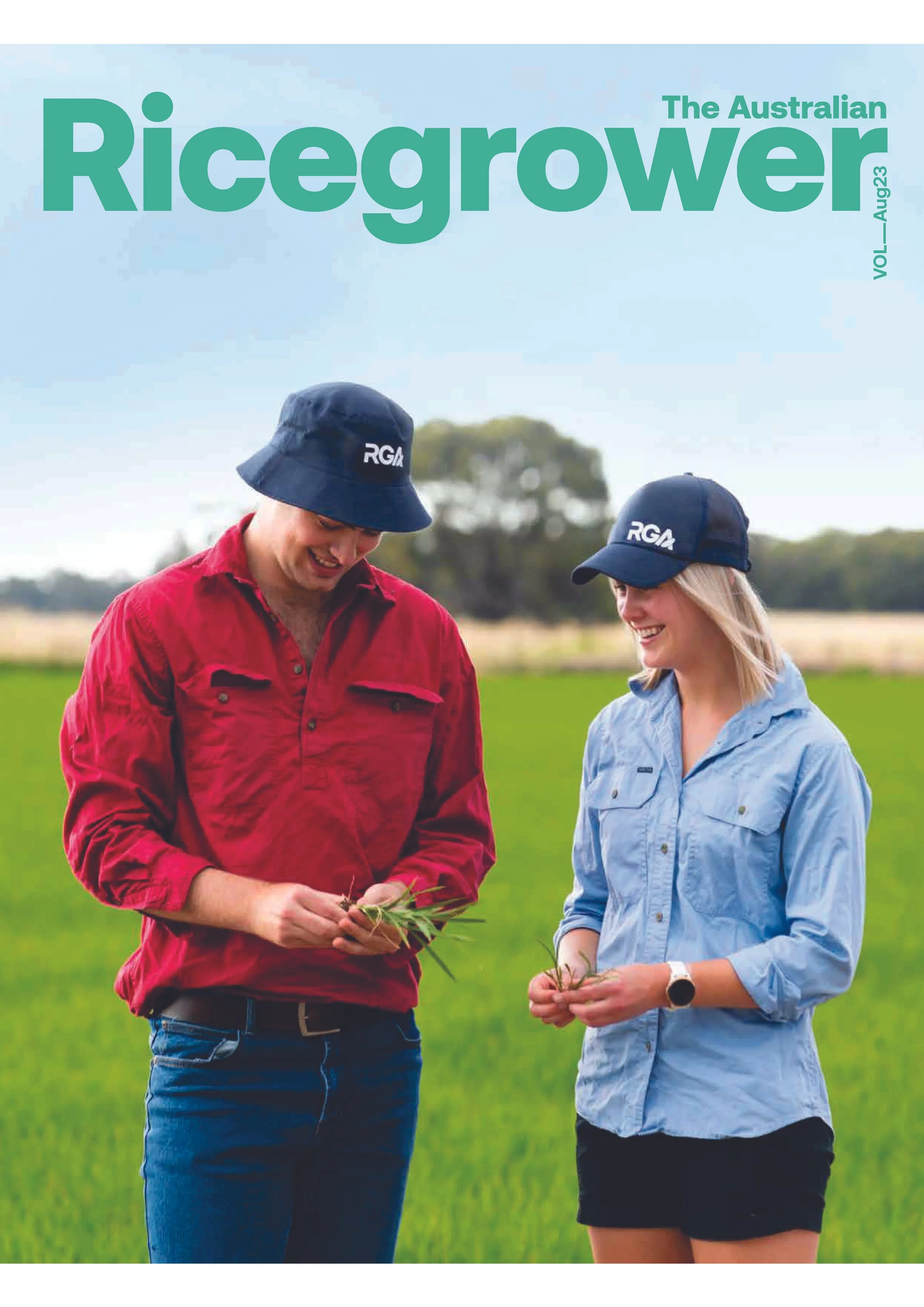 Two people standing in a green field, smiling and holding plants, wearing caps with 'RGA' on them, one in a red shirt and the other in a light blue shirt.