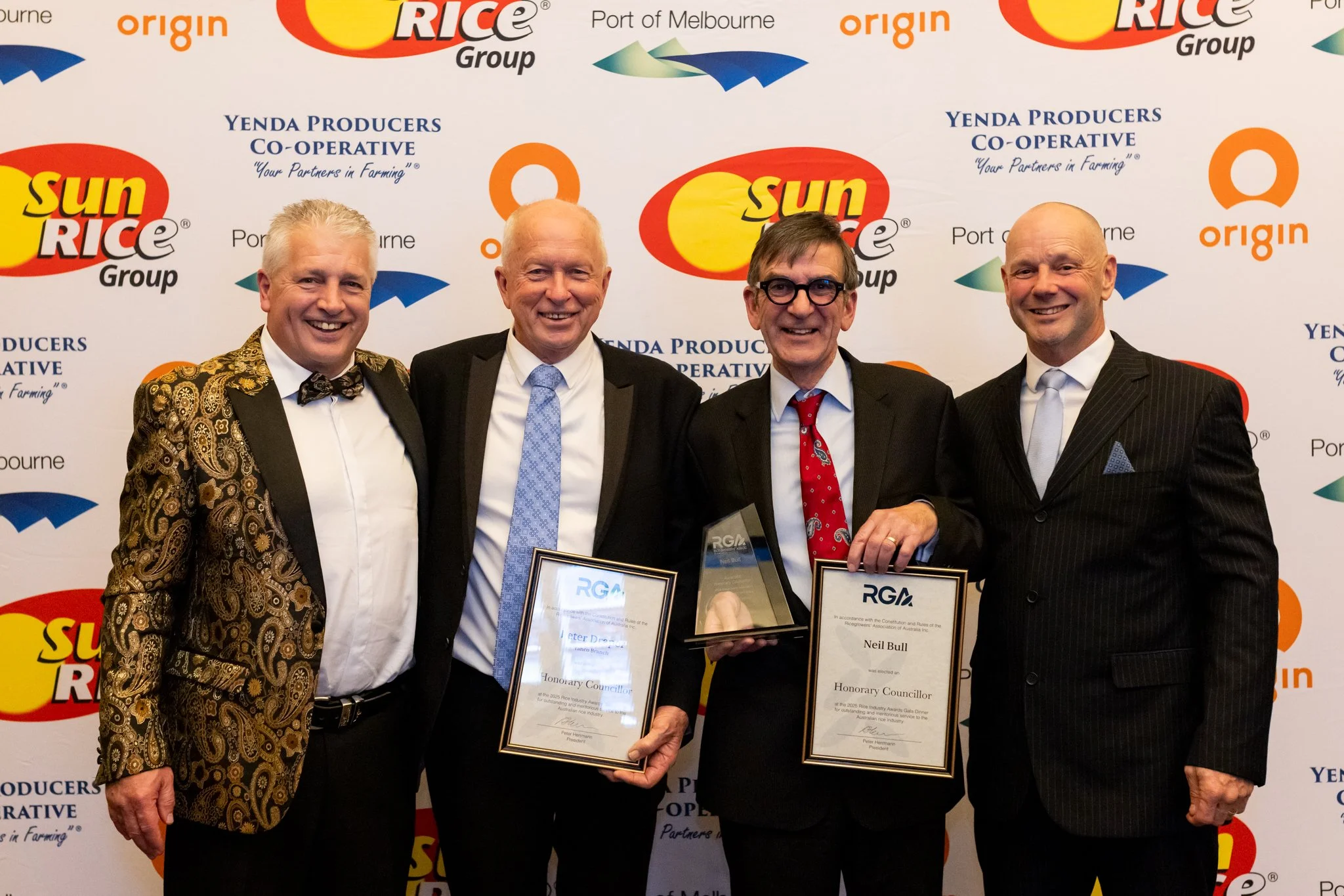 Four men in formal suits standing together at an awards ceremony, holding framed certificates and a trophy, with a backdrop featuring logos such as SunRice Group, Origin, and Yenda Producers Co-operative.