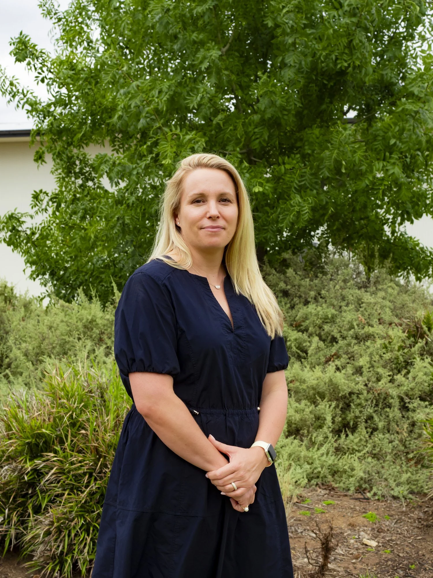 A woman with blonde hair smiling, wearing a colorful, patterned dress, standing against a plain white wall.