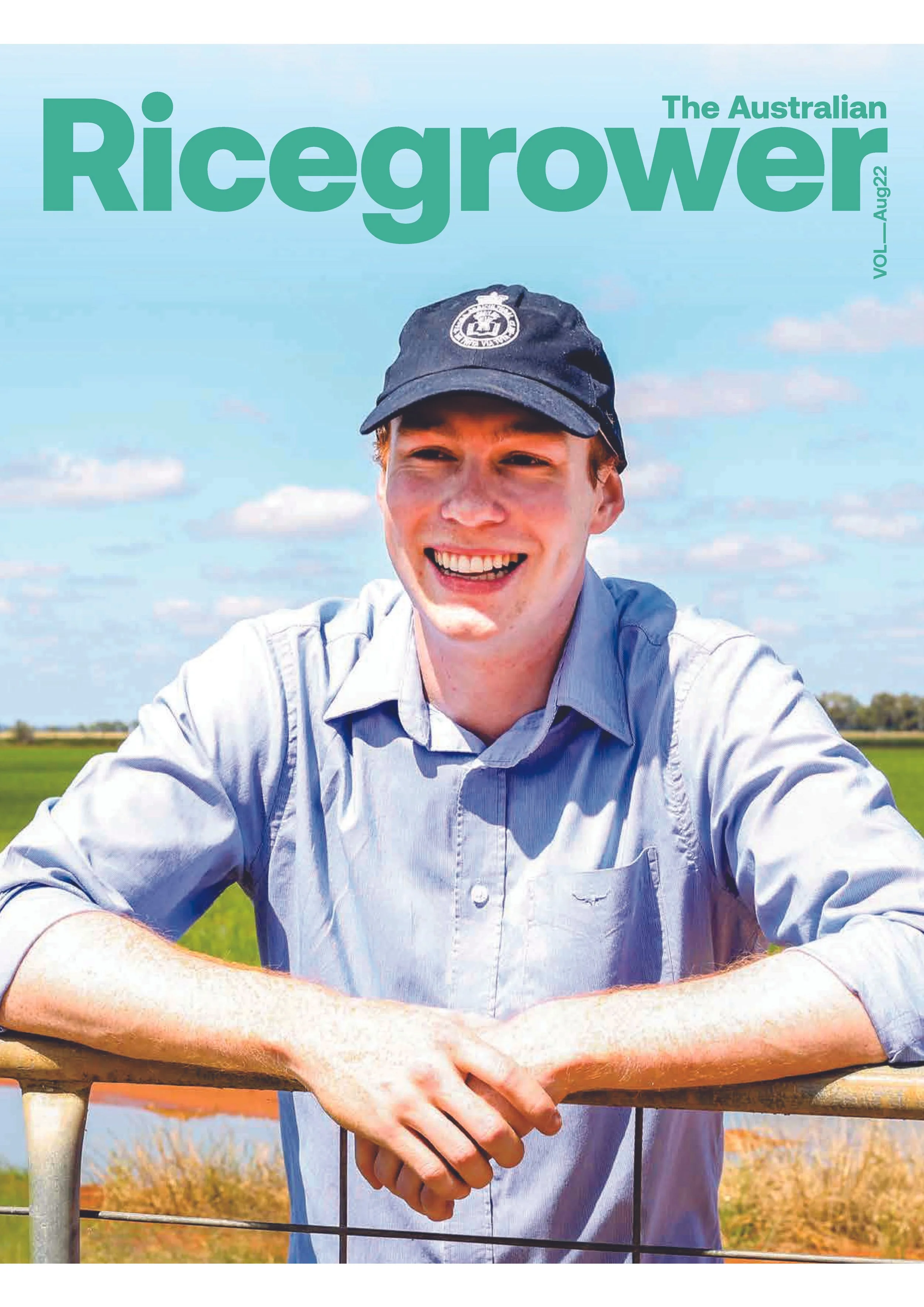 Cover image of Ricegrower magazine featuring a young man smiling outdoors in a field, wearing a blue shirt and a dark cap with a badge, with a blue sky and scattered clouds in the background.