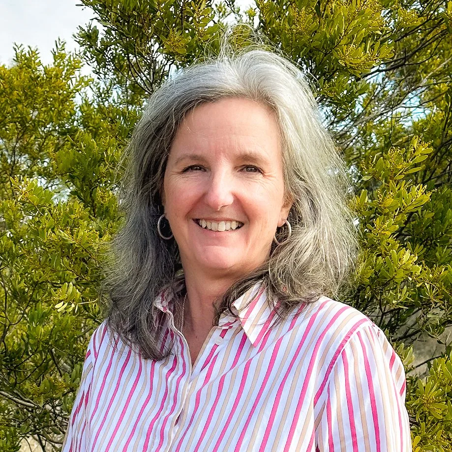 A woman with gray hair, wearing a striped pink and white shirt, standing outdoors in front of green shrubbery, smiling at the camera.