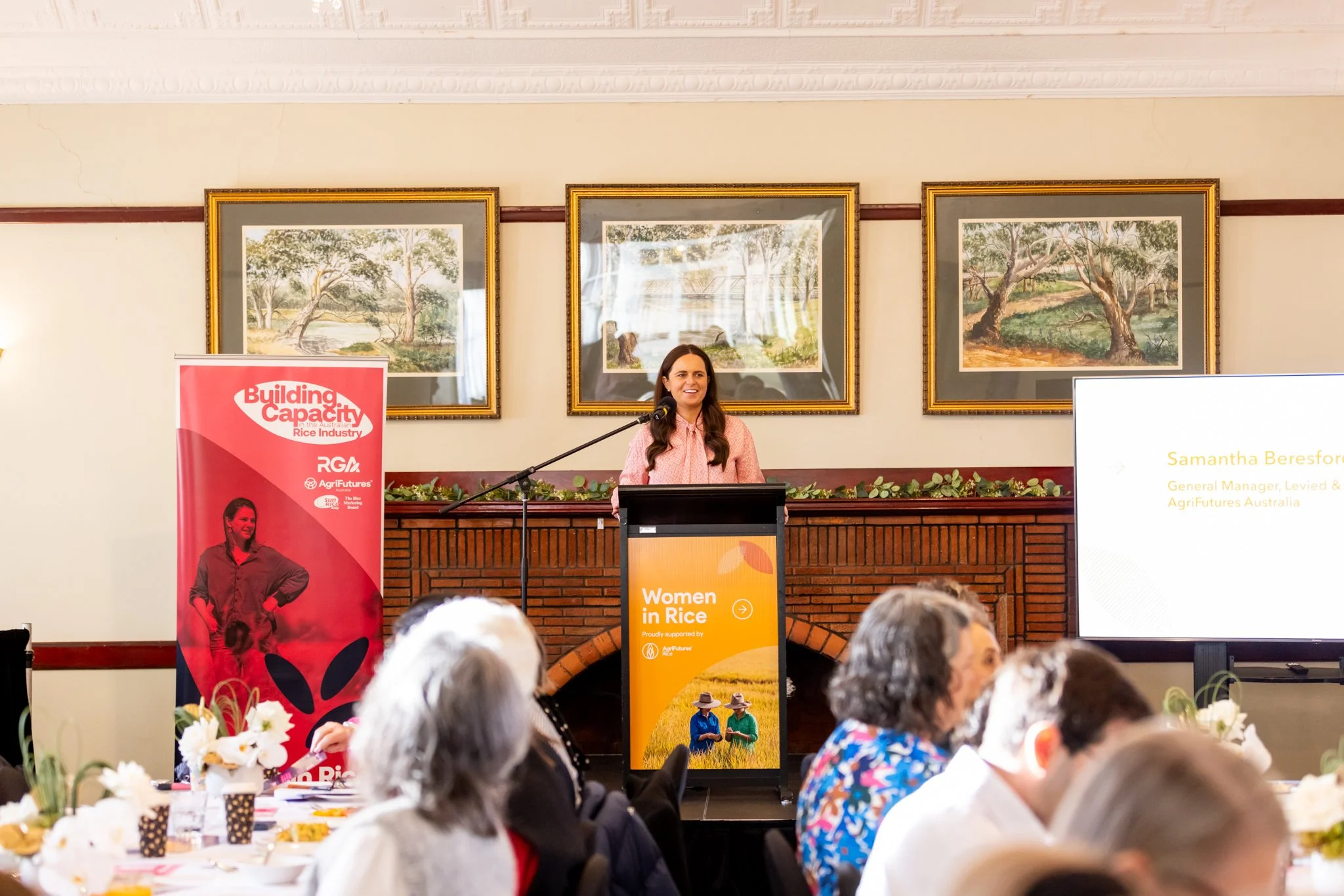 A woman speaking at a podium during a conference with a sign that reads 'Women in Rice' and a screen showing her name and title, with several papers, decorations, and attendees in the foreground.
