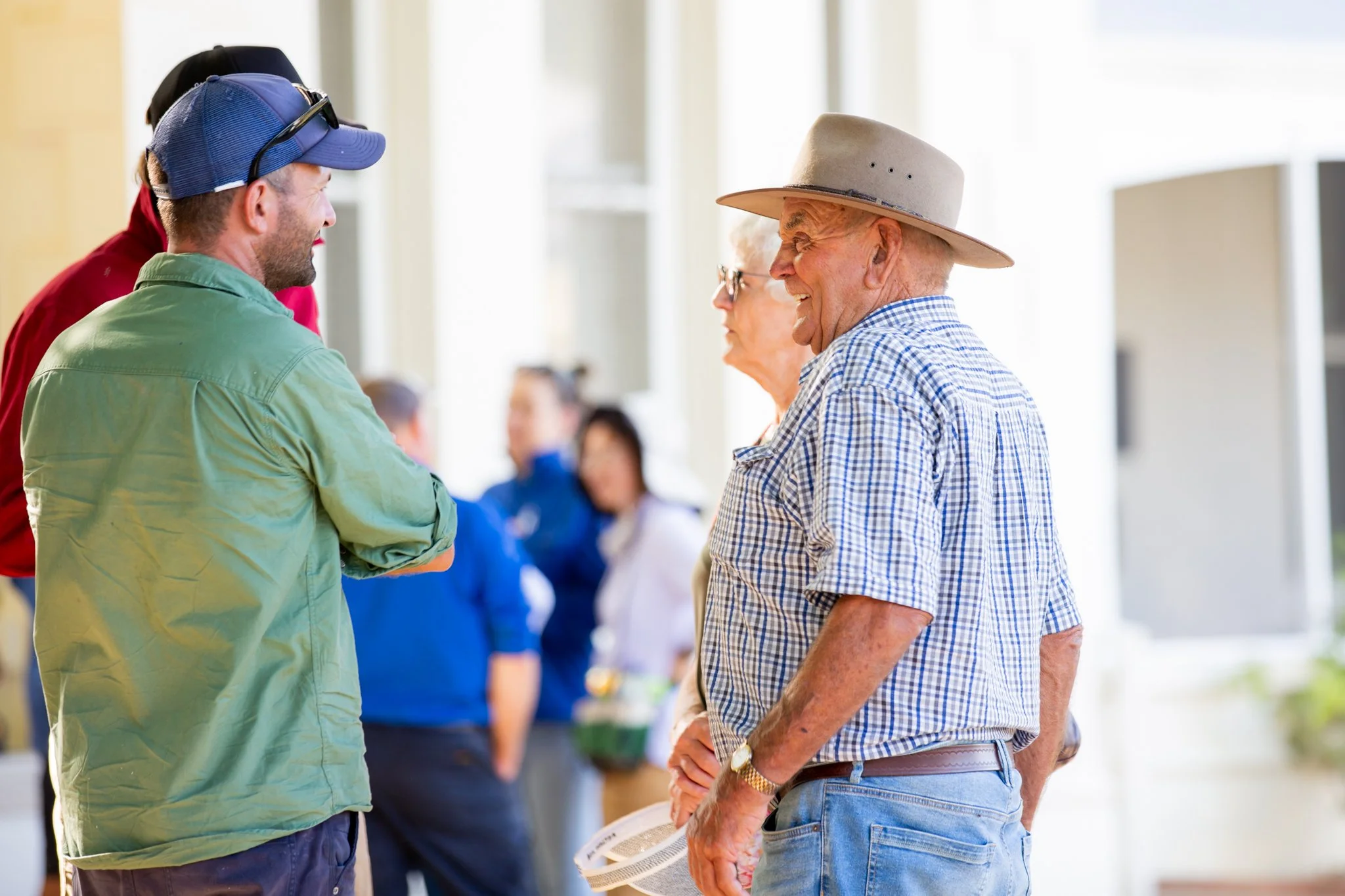 Older man in a cowboy hat and plaid shirt smiling and talking with a younger man in a green jacket and baseball cap at a social event.