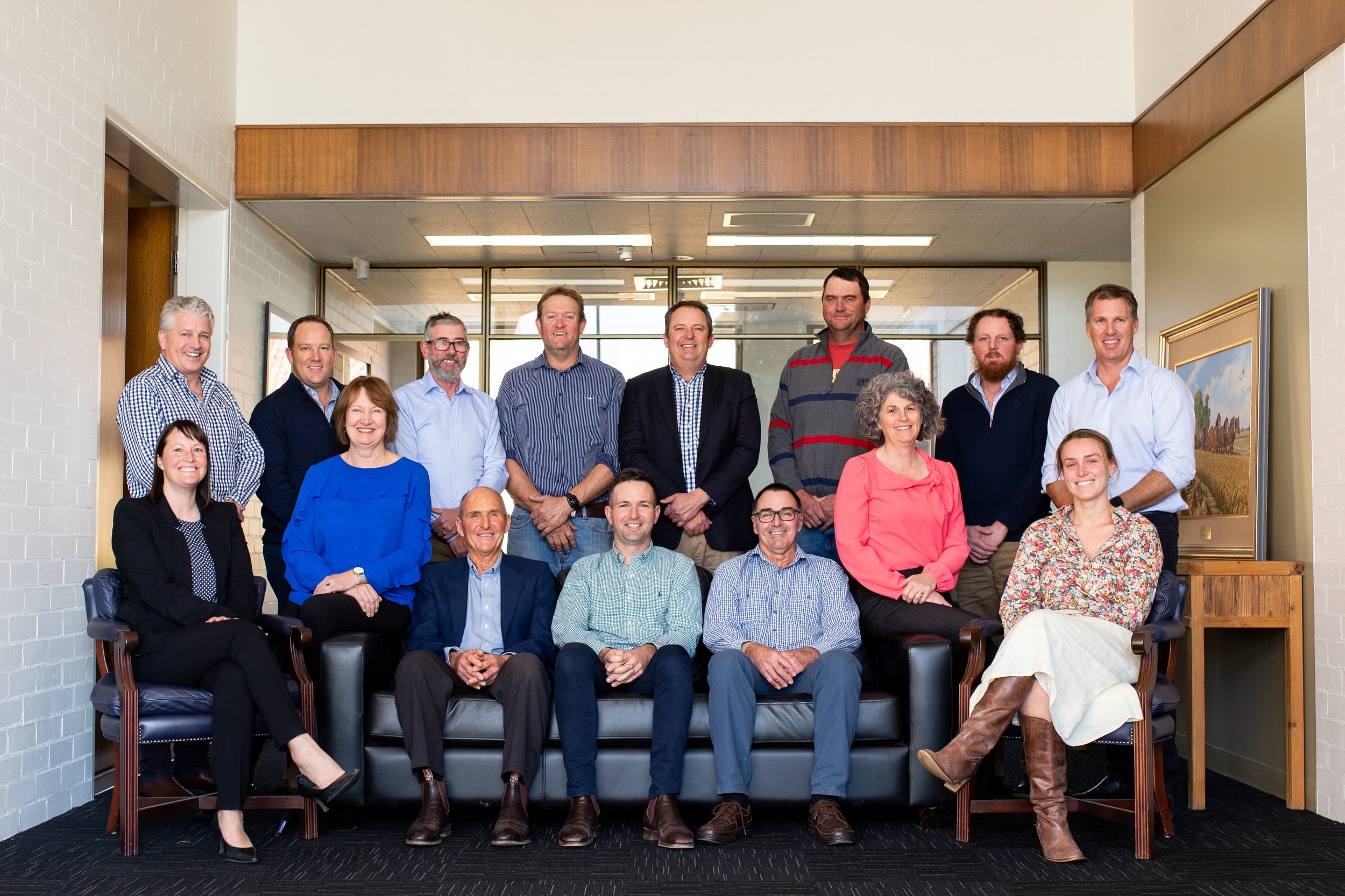 Group photo of fifteen people, thirteen men and two women, standing and sitting in a corporate office setting with artwork and glass walls.