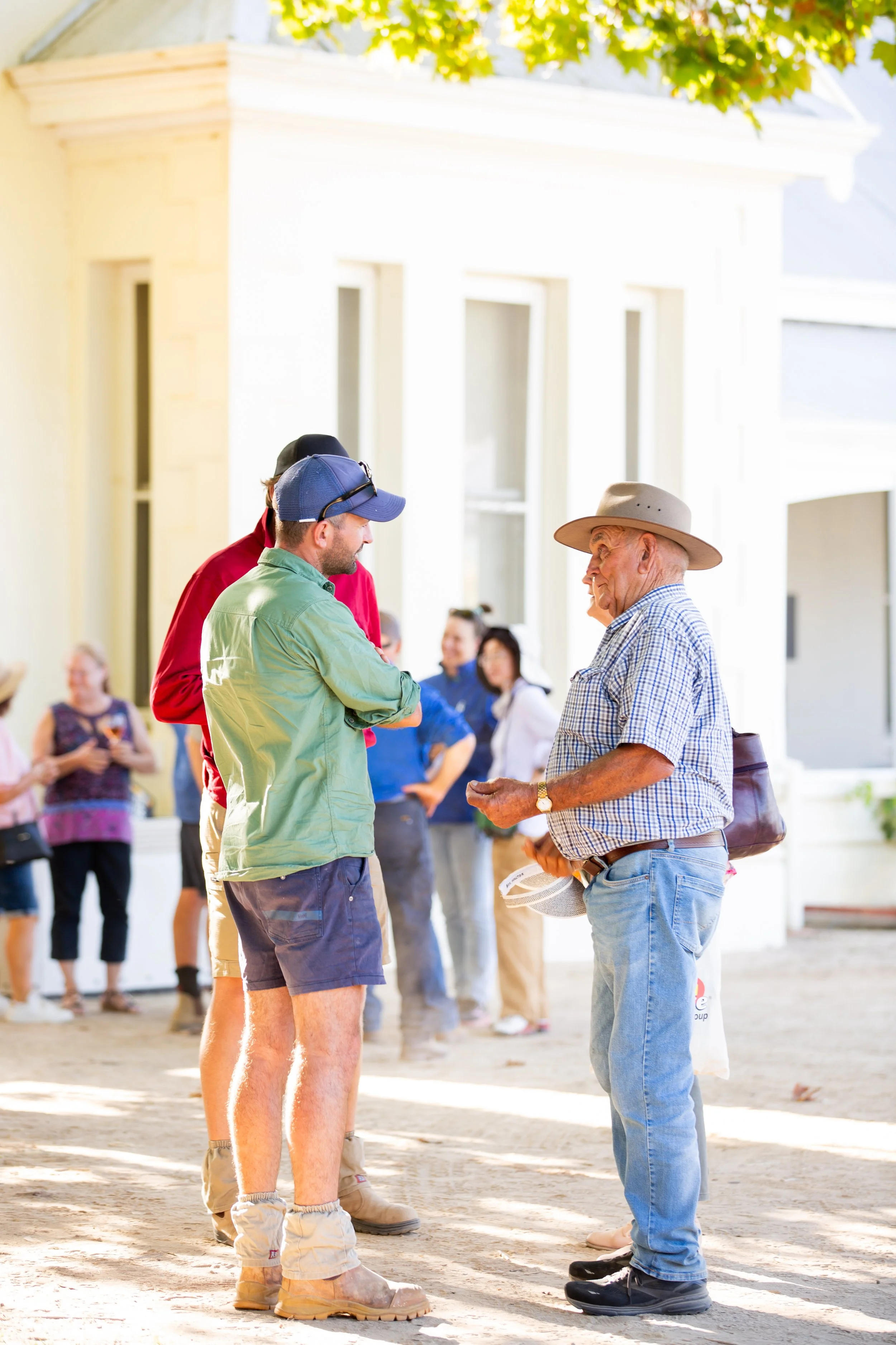Older man in a plaid shirt and wide-brimmed hat talking to two younger men, with a group of people standing in the background outside a white building on a sunny day.