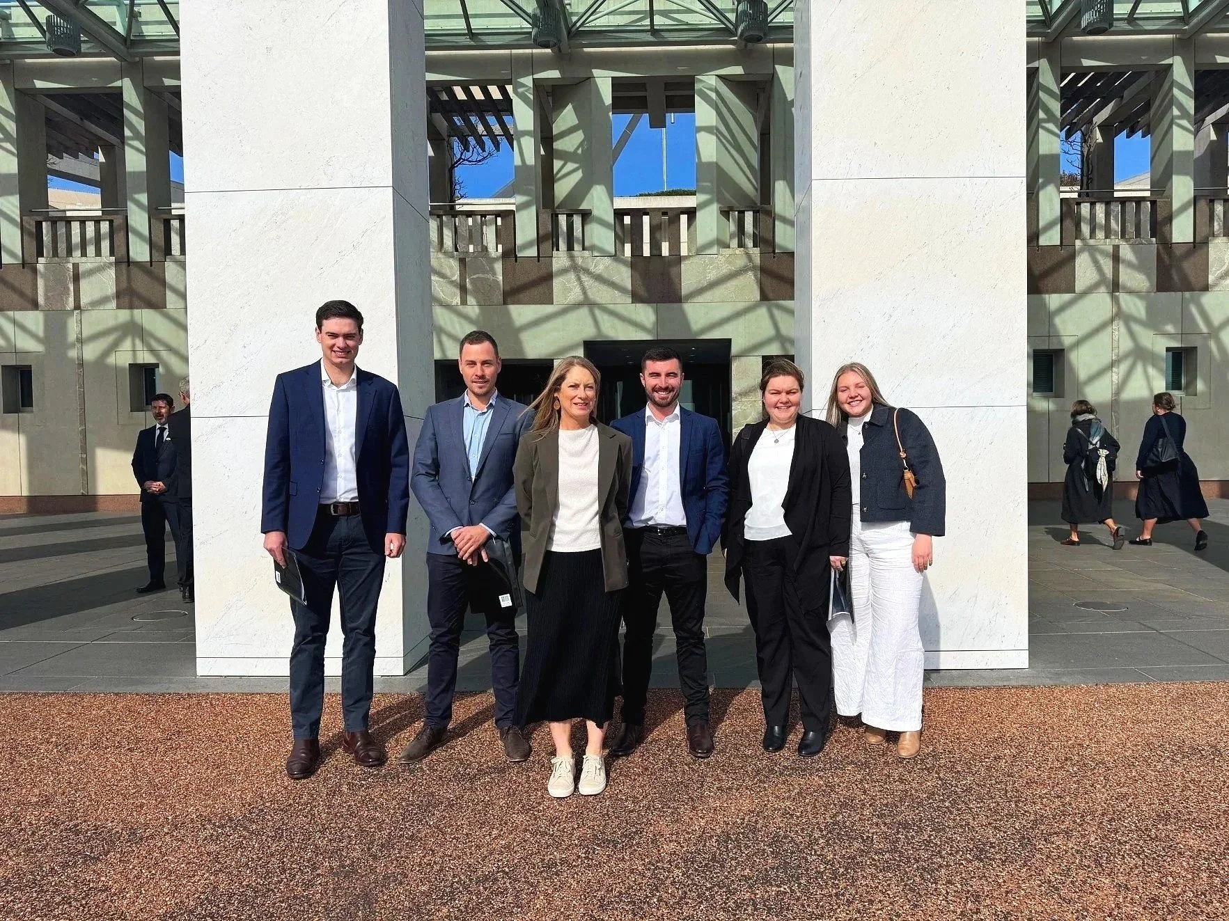 Group of seven professionally dressed people standing together outside in front of a large modern building on a sunny day, smiling for the camera.