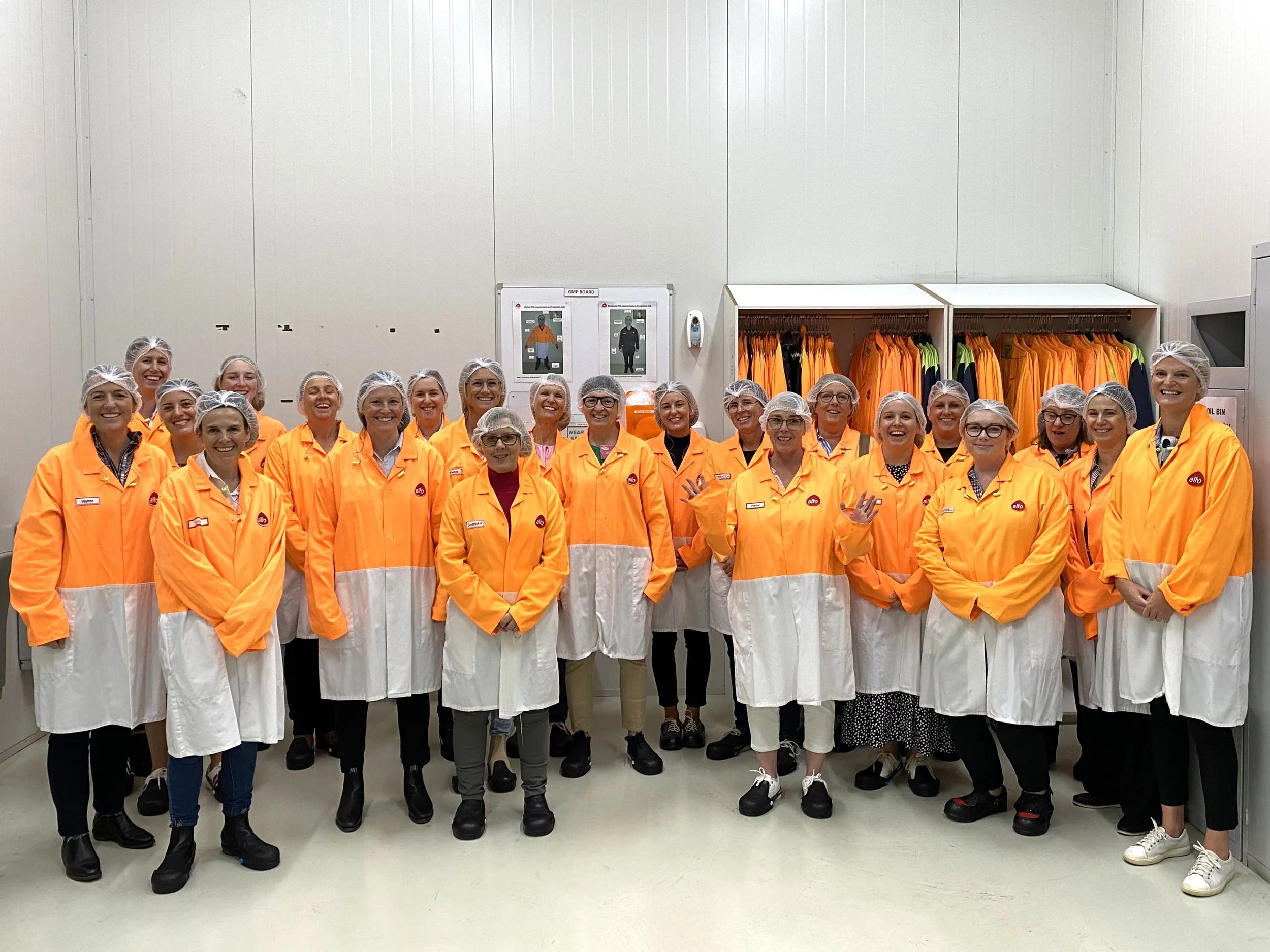 Group of women wearing orange and white uniforms with hair nets, standing in a room with white walls and clothing racks with orange uniforms.