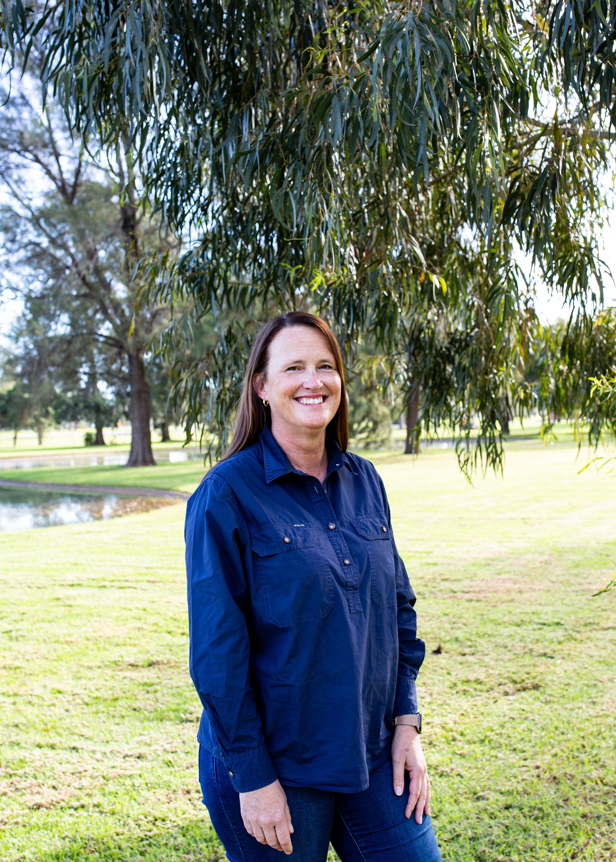 Smiling woman standing outdoors in a park with green grass, trees, and a pond in the background, wearing a blue shirt.