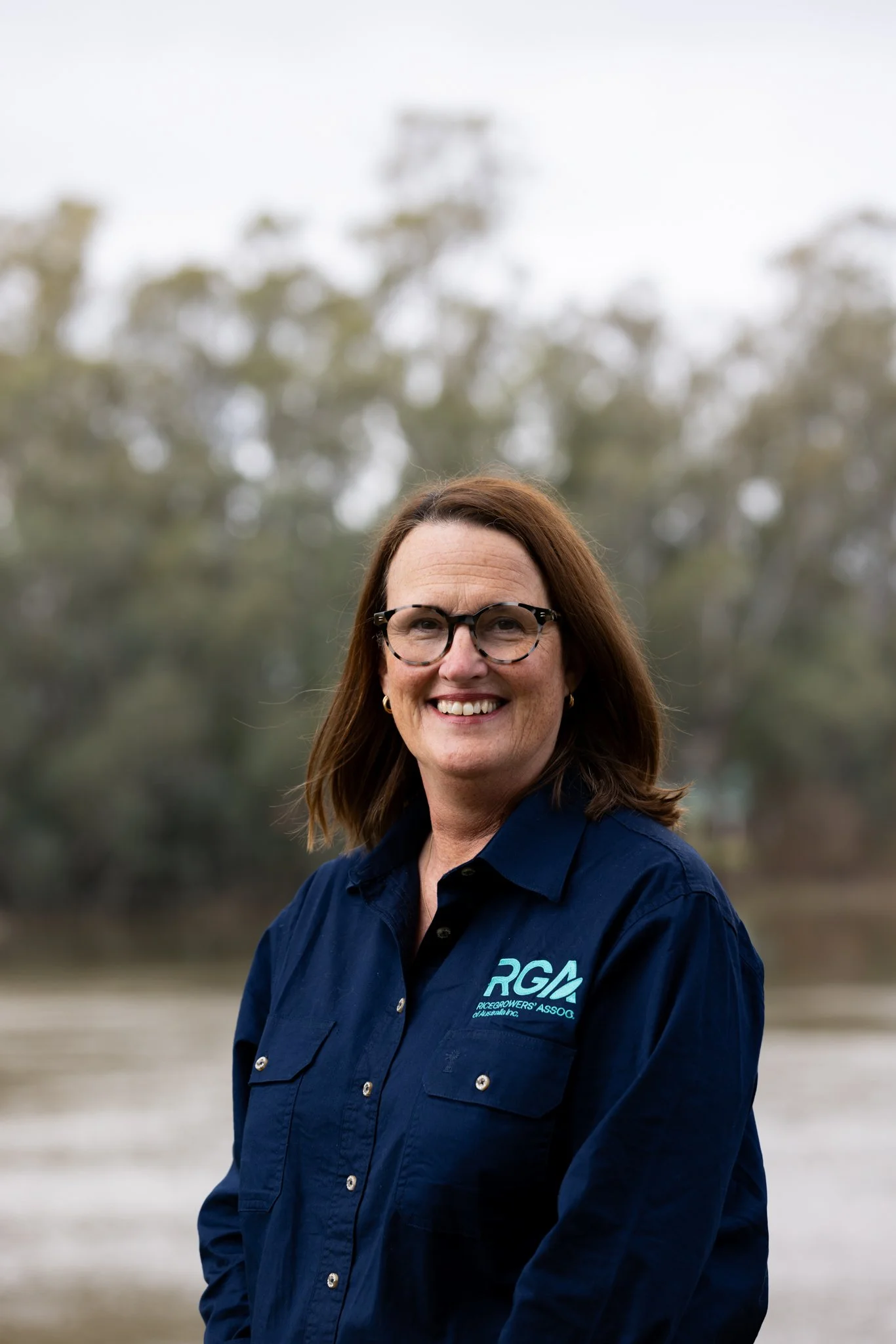 A woman is standing outdoors in a park with trees and a pond in the background. She has long brown hair, is smiling, and is wearing a blue button-up shirt and jeans.