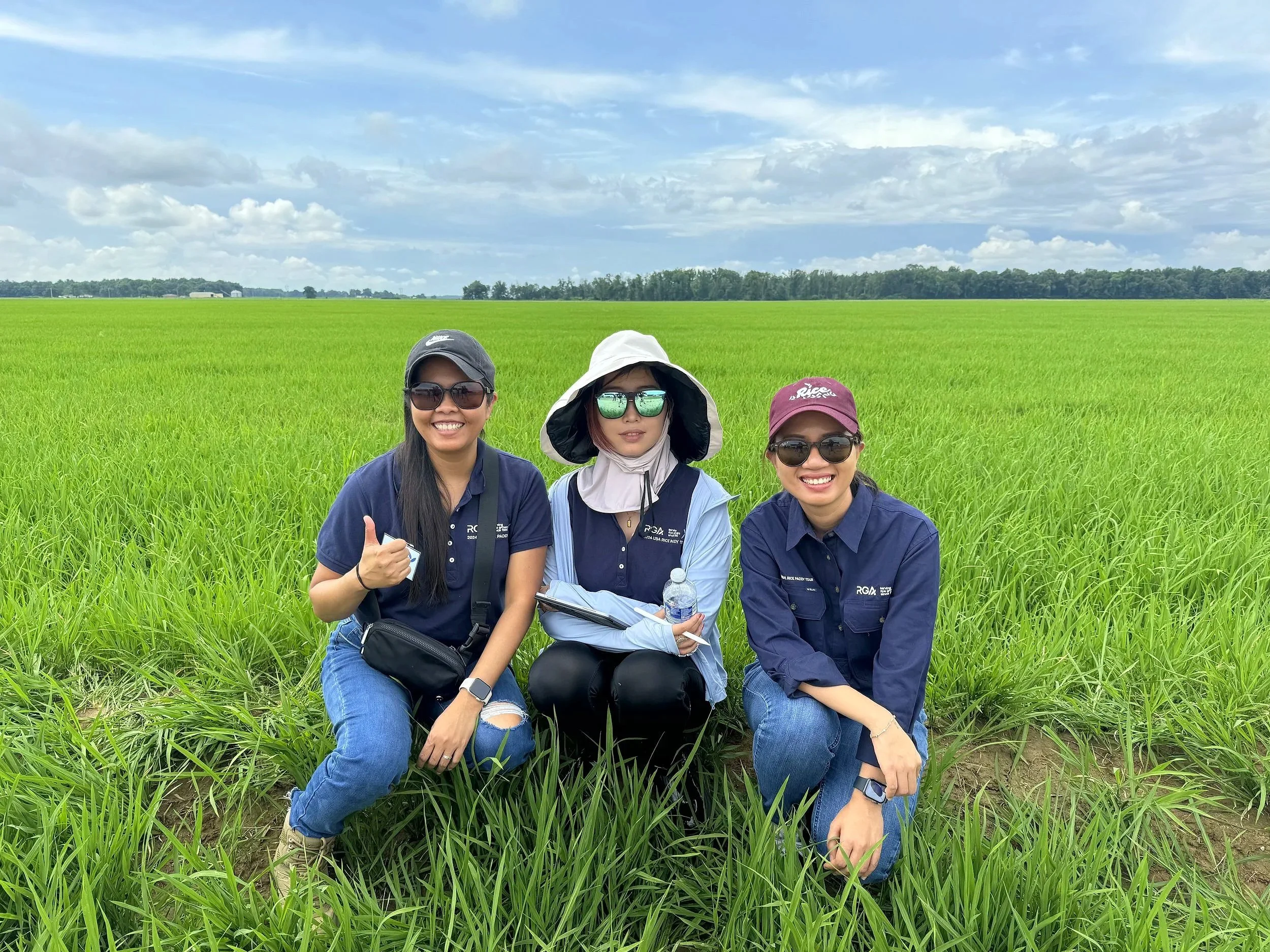 Three women in sunglasses and casual clothing kneeling in a green rice field with a cloudy sky in the background.