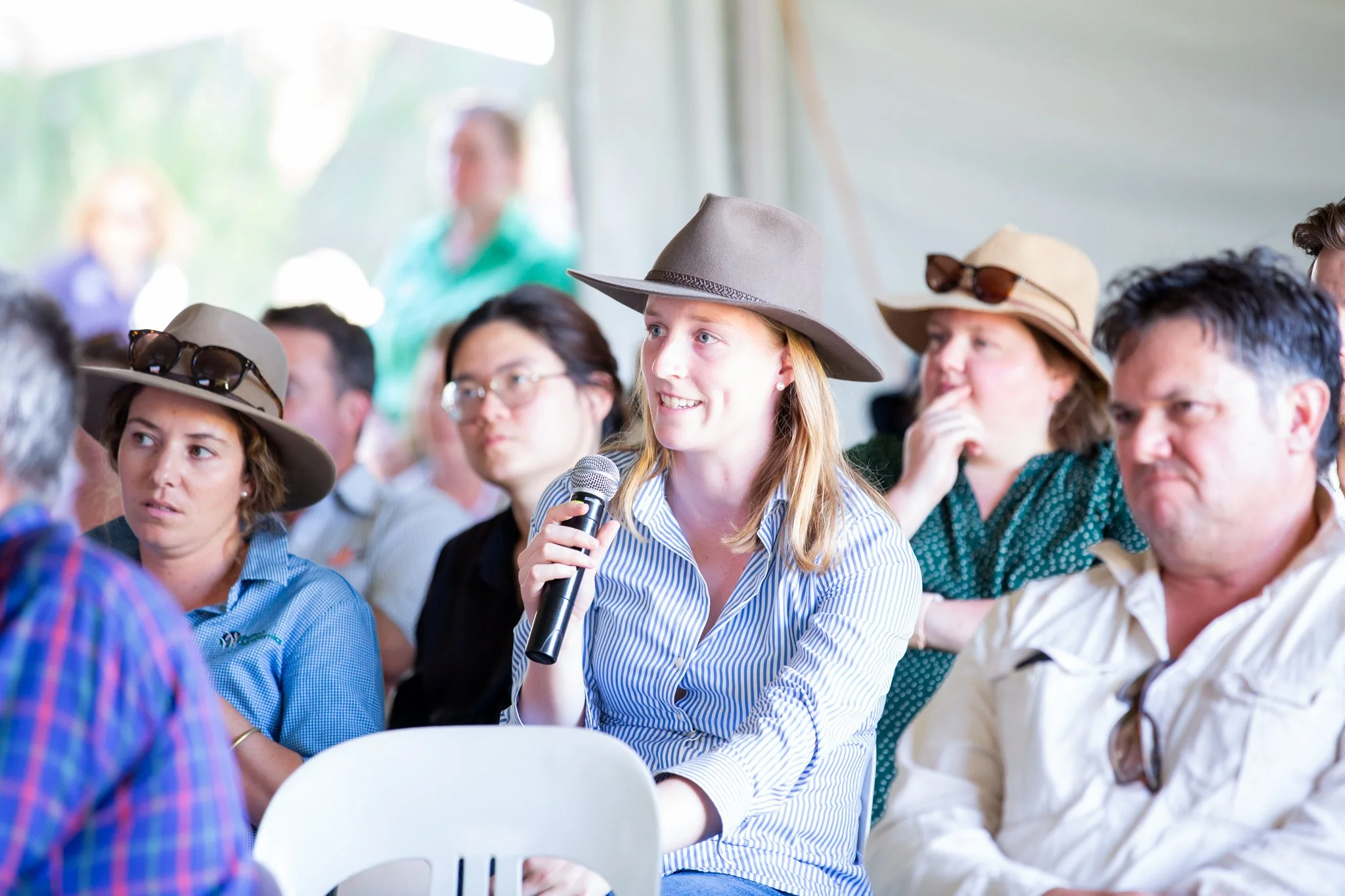 A woman with red hair wearing a gray hat, holding a microphone, sitting in an audience at an indoor event.