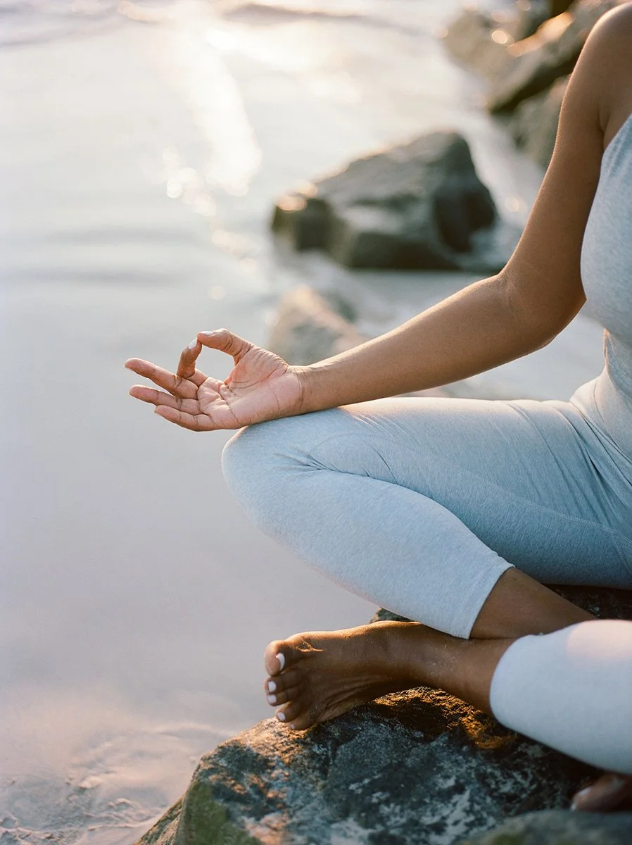 Person practicing yoga or meditation on a beach, sitting cross-legged on a rock with a hand in the Gyan mudra, near the water at sunset.
