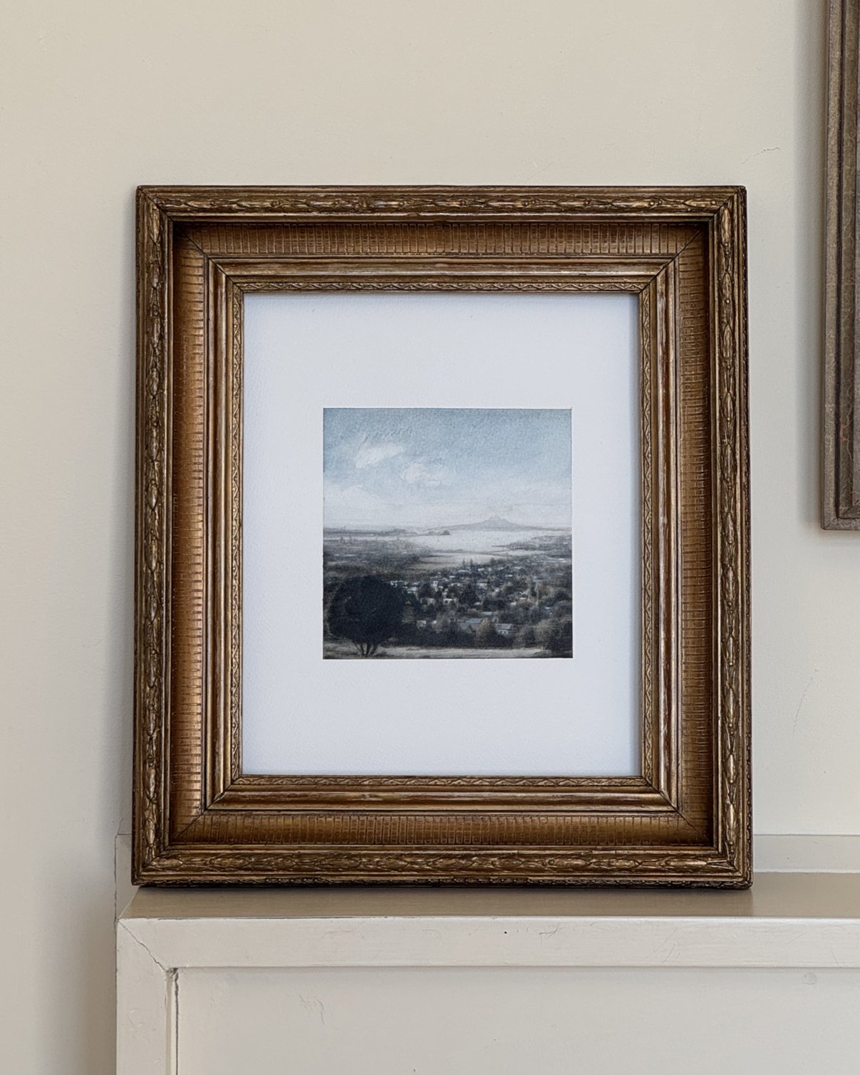 Framed landscape painting of Rangitoto scene with hills, trees, and a cloudy sky, displayed on a light-colored wall.