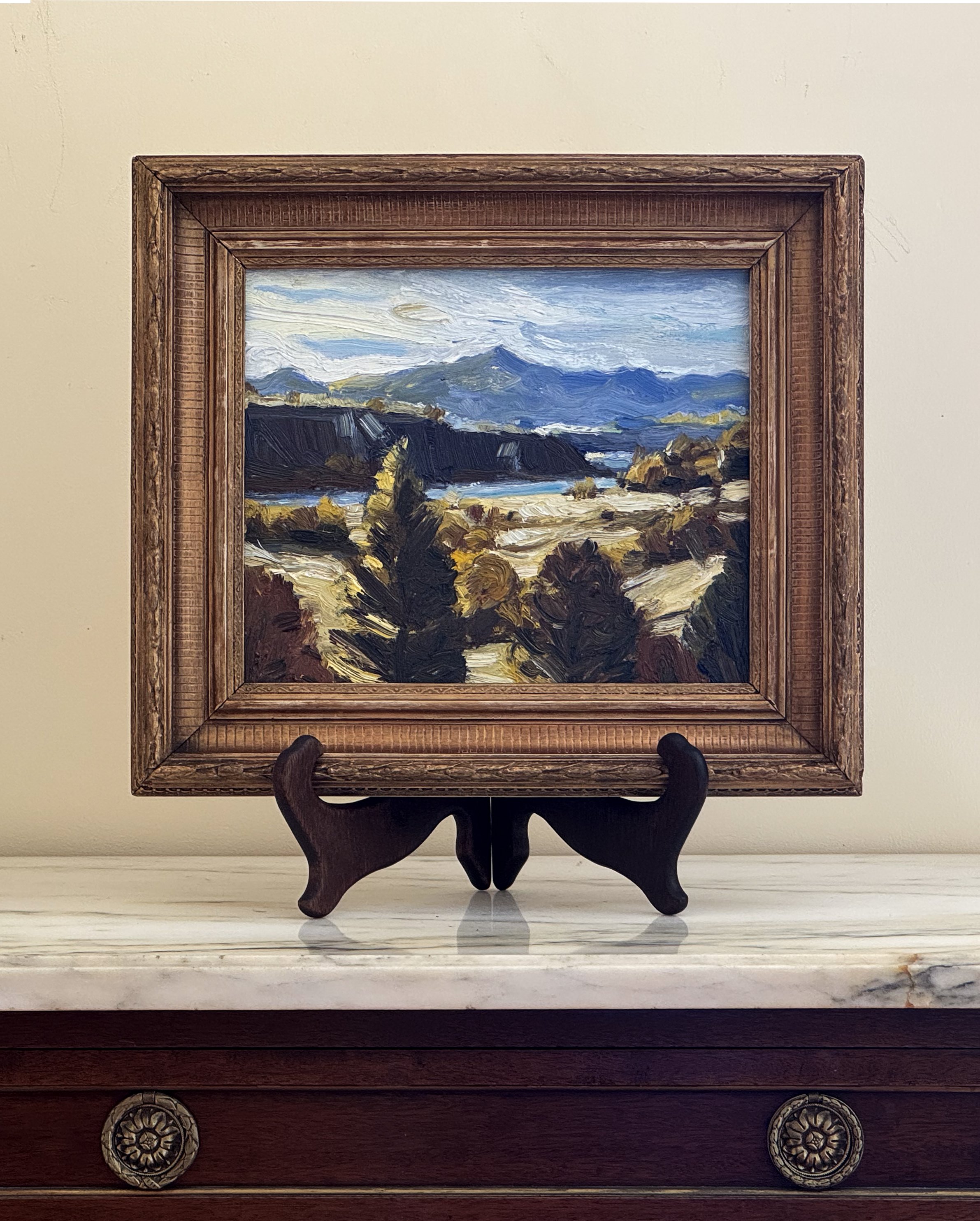 Central Otago landscape painting of mountains, trees, and a river under a partly cloudy sky, displayed on a wooden stand on a marble surface.