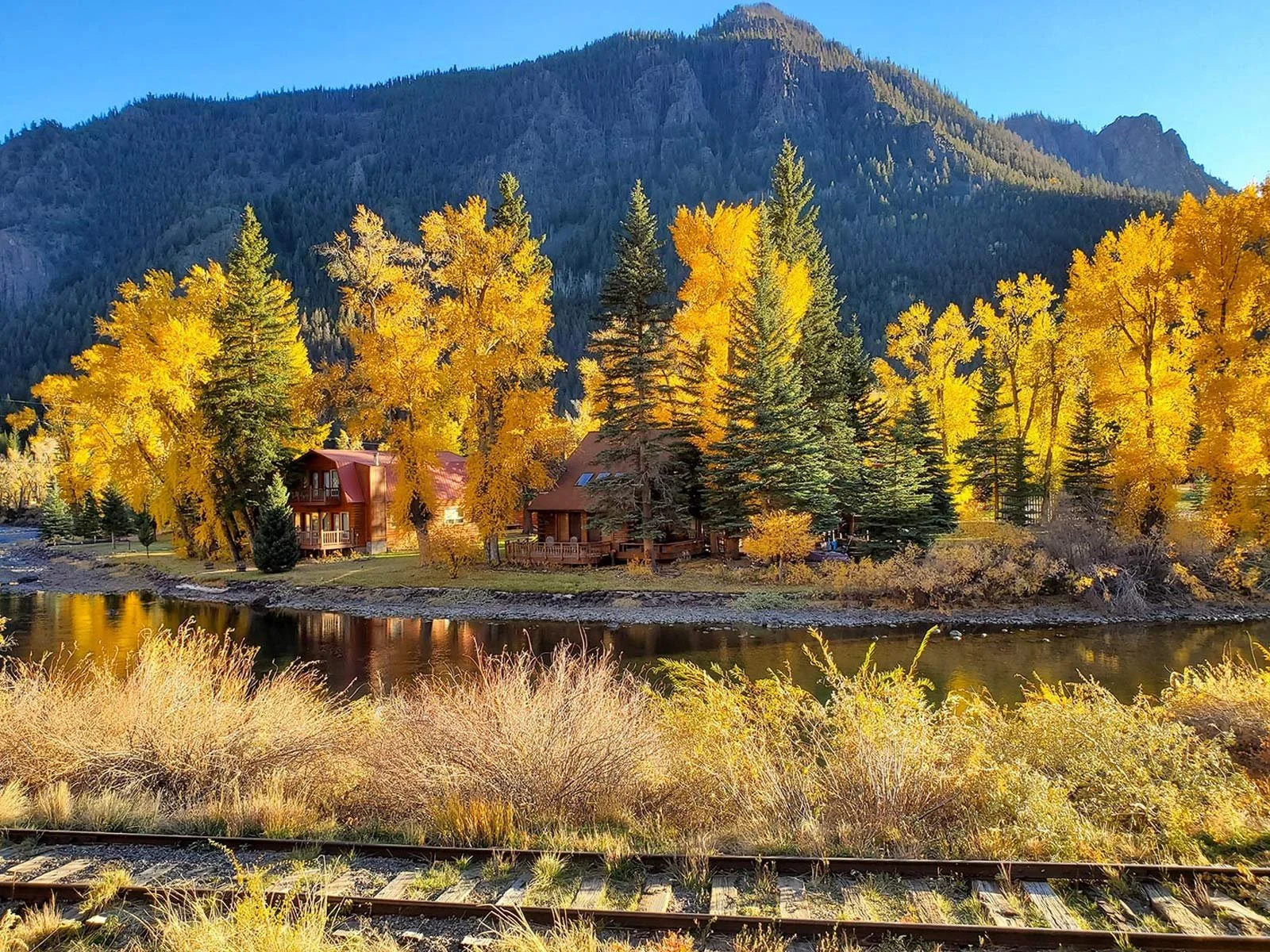 Scenic autumn landscape in Creede, Colorado featuring vibrant yellow aspen trees reflecting in a calm river with mountain peaks in the background.