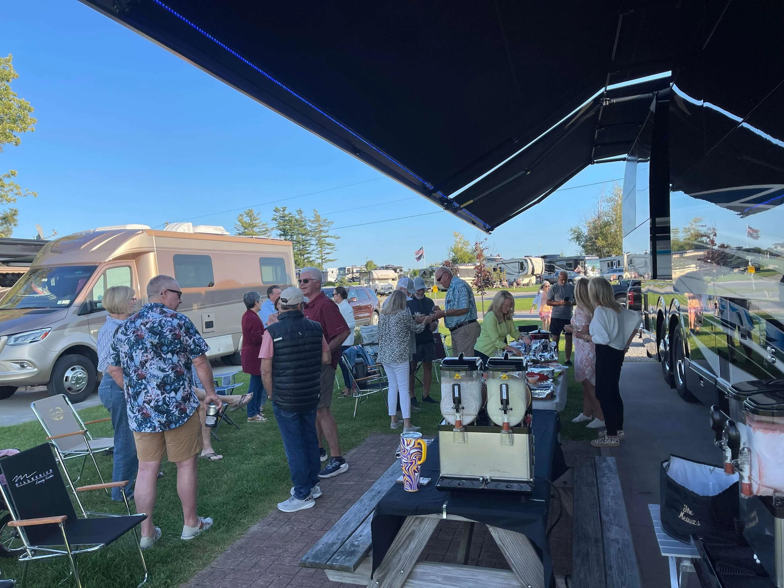 Members enjoying an outdoor gathering near luxury RVs, with food and drinks set up under a canopy.
