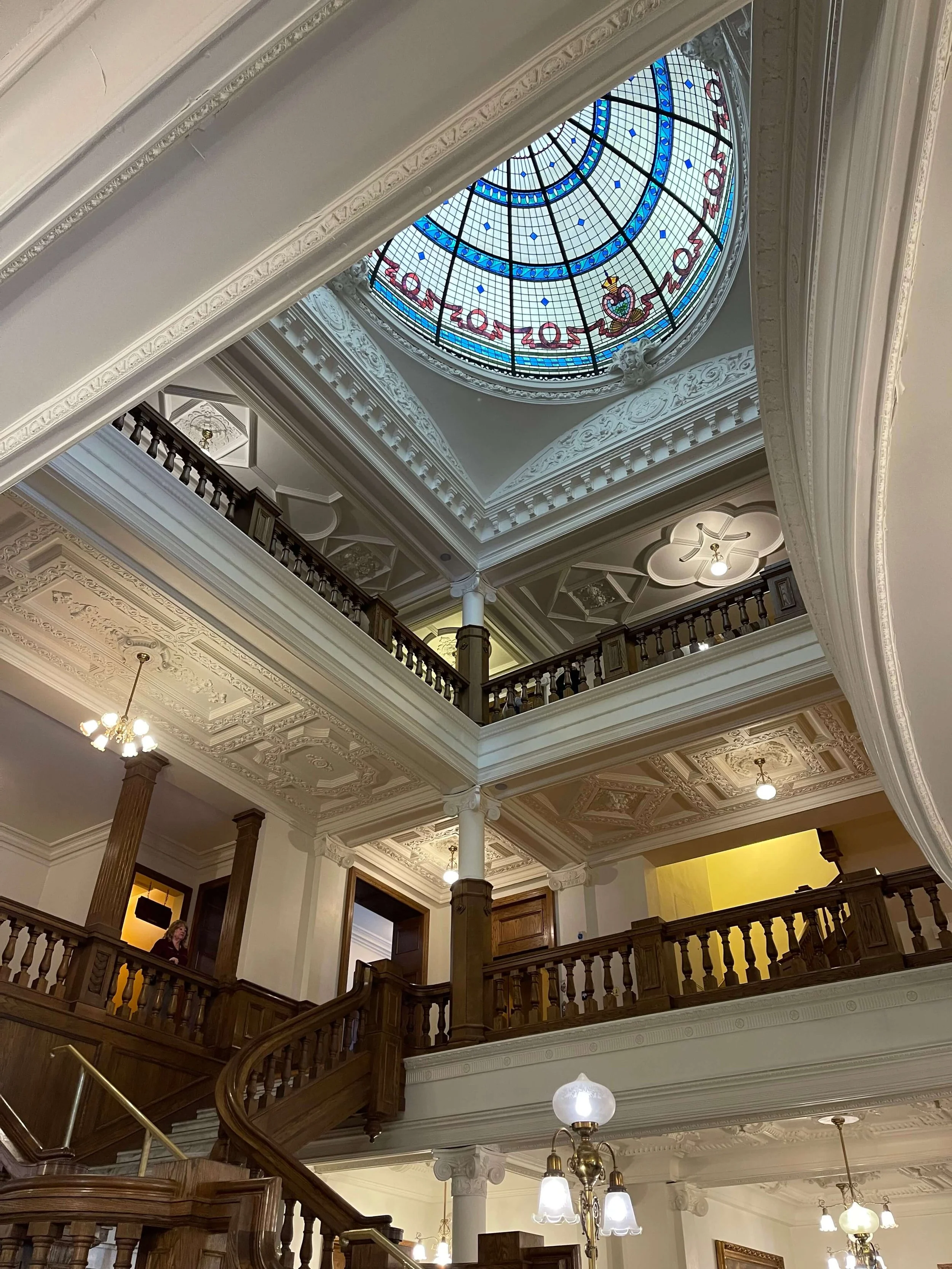 Ornate interior of a historic building with a stained glass ceiling and grand wooden staircase.
