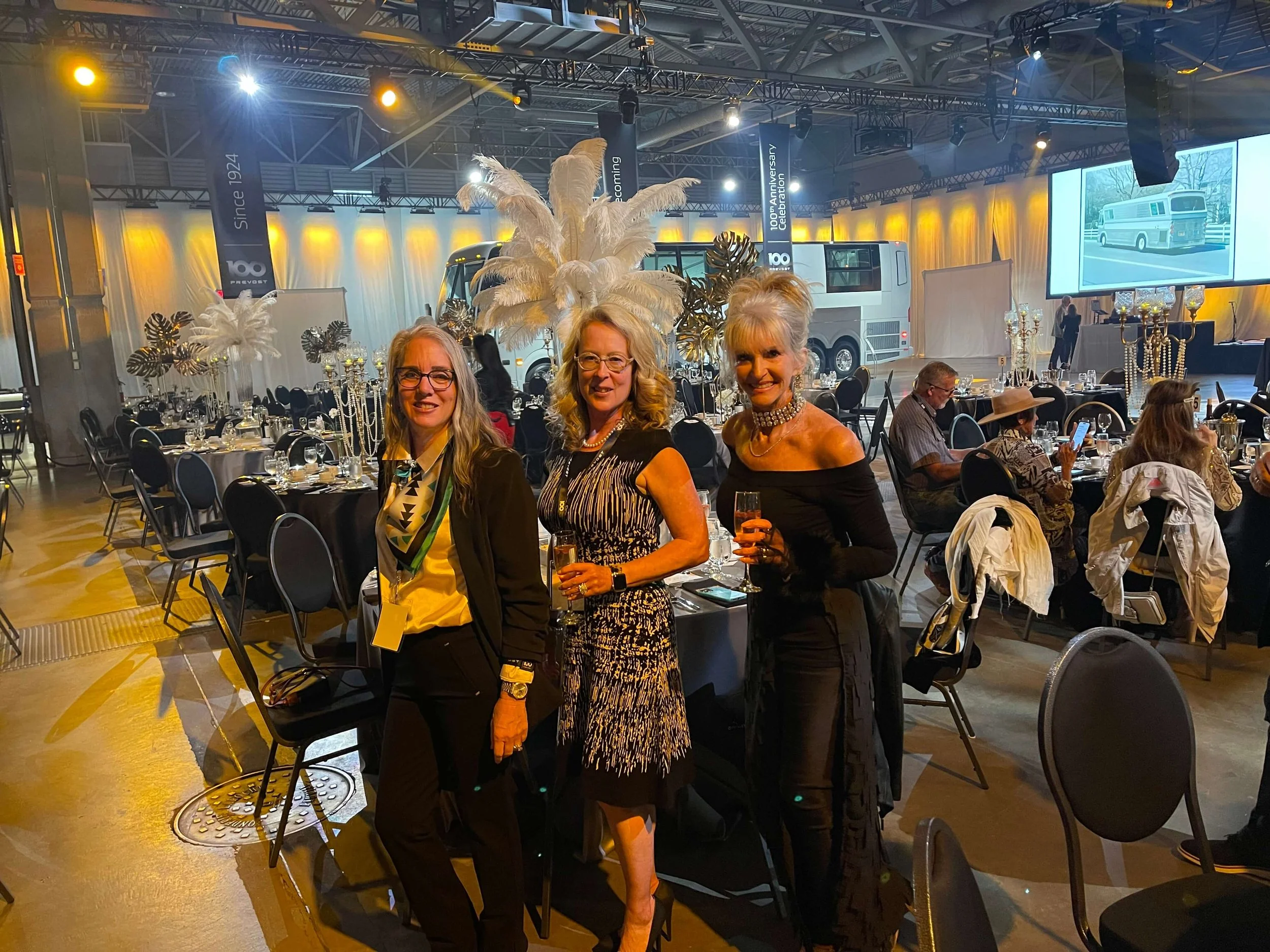 Three women smiling at a gala event with elaborate table settings and feathered decorations.