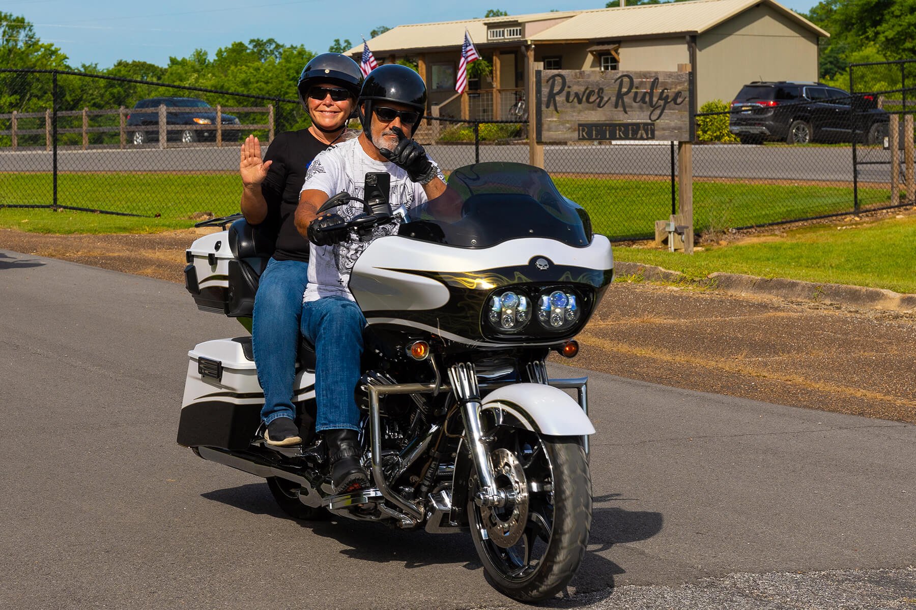 Two club members on a white motorcycle, waving and smiling as they ride past the River Ridge Retreat sign.