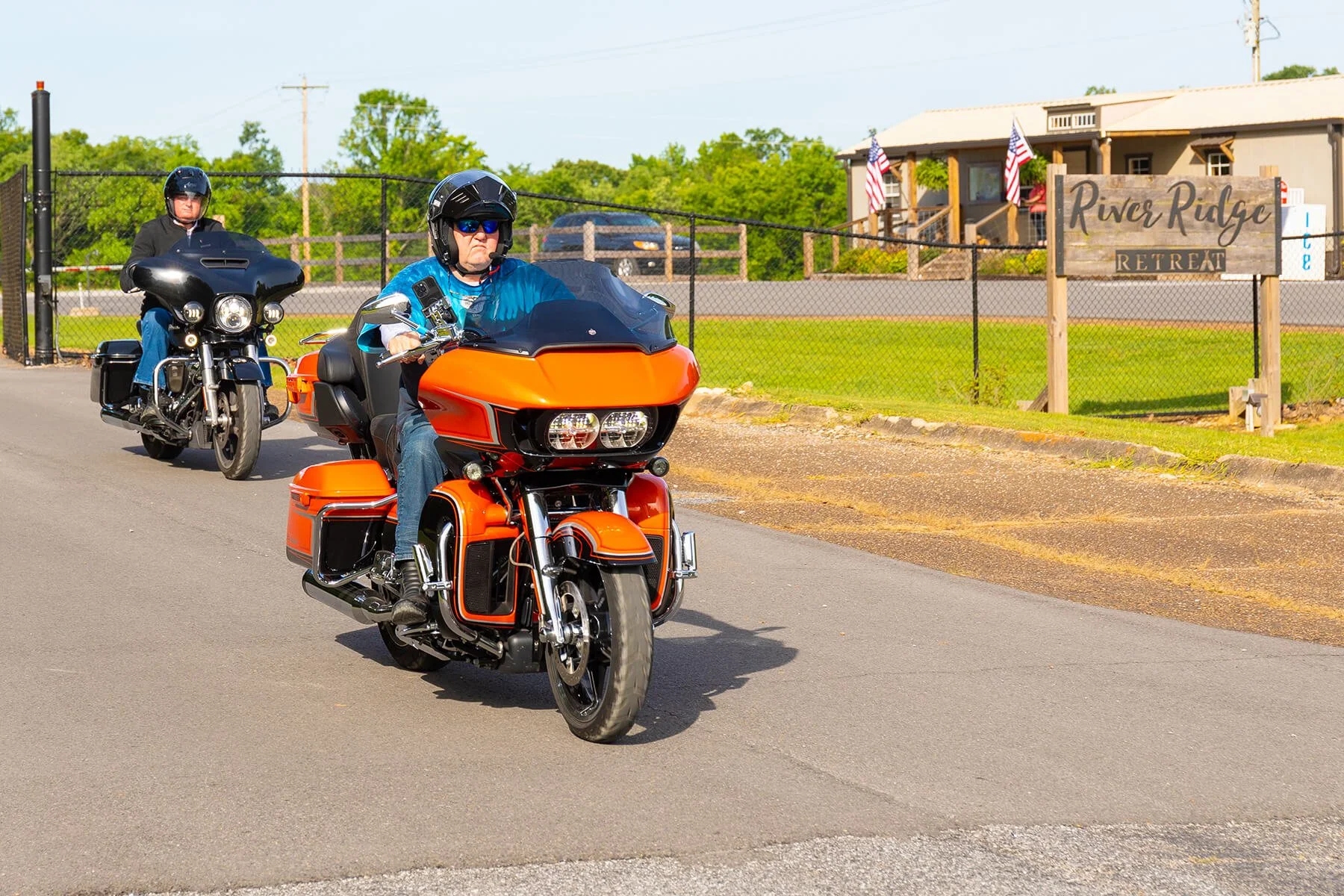 Bikers riding Harley-Davidson motorcycles past the River Ridge Retreat, led by a rider on an orange trike. 