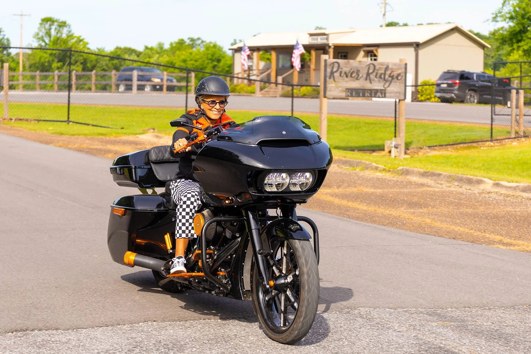 Female motorcyclist in a black and orange jacket and checkered pants, riding a black trike past the River Ridge Retreat sign. 