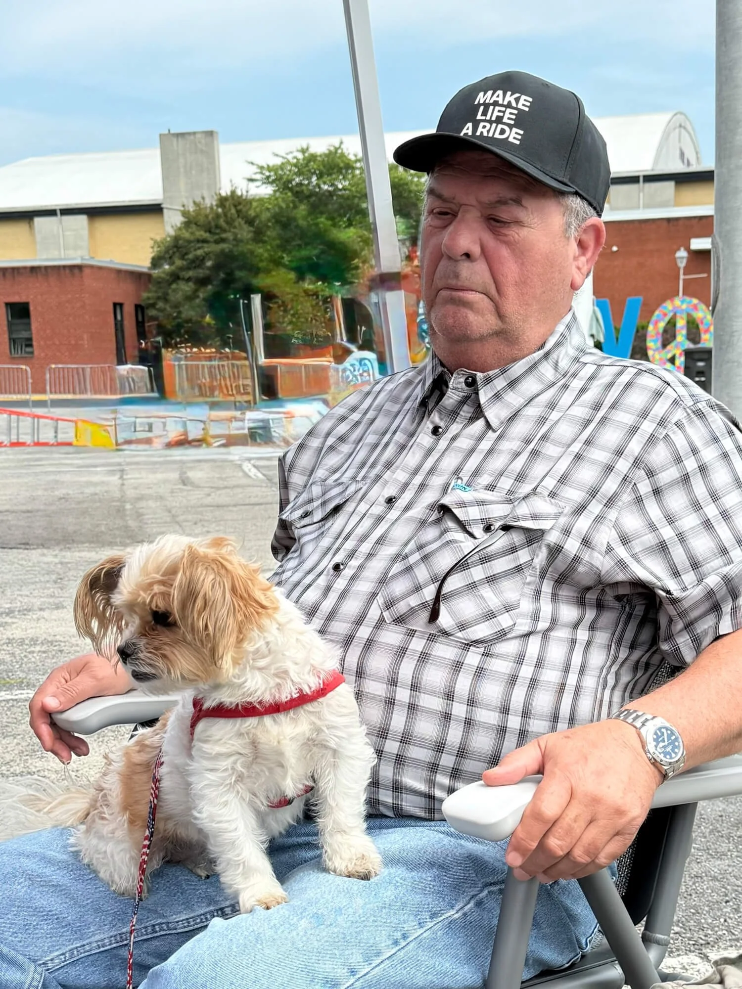 A man wearing a "MAKE LIFE A RIDE" hat sitting down with a small dog on his lap.