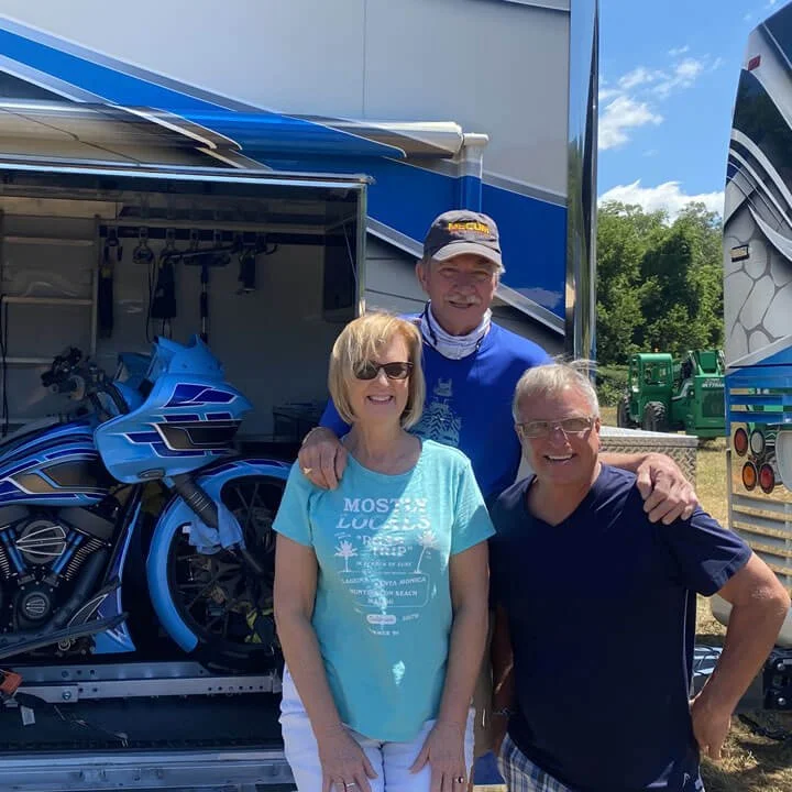 Three people posing in front of an open RV trailer bay with a custom motorcycle.