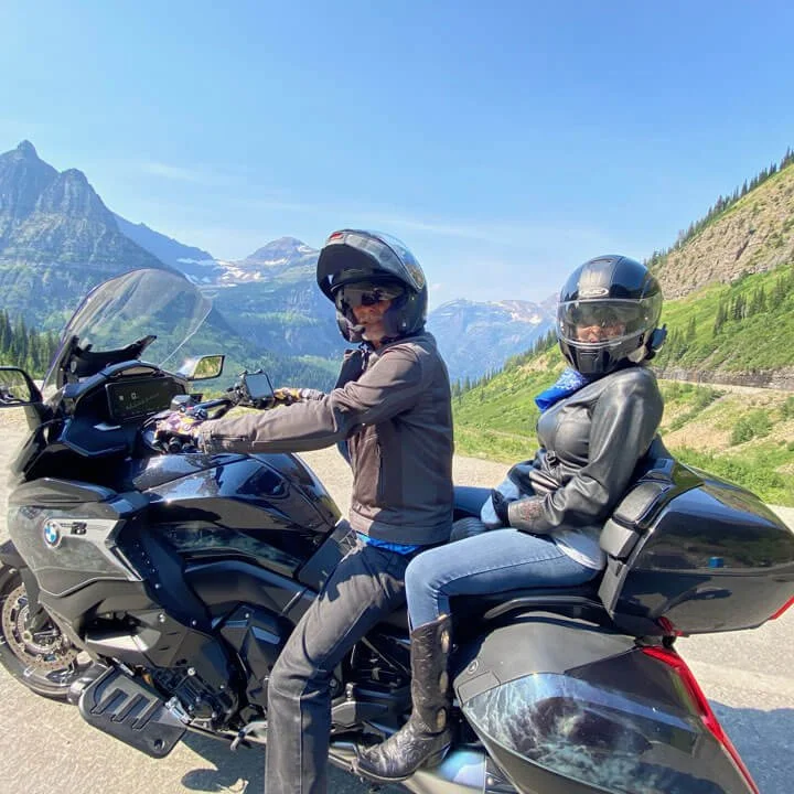 Two riders on a black BMW motorcycle with mountains in the background.