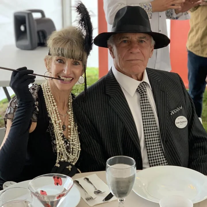 Couple in 1920s flapper and gangster costumes at themed party with pearls, feather headpiece, and pinstripe suit.