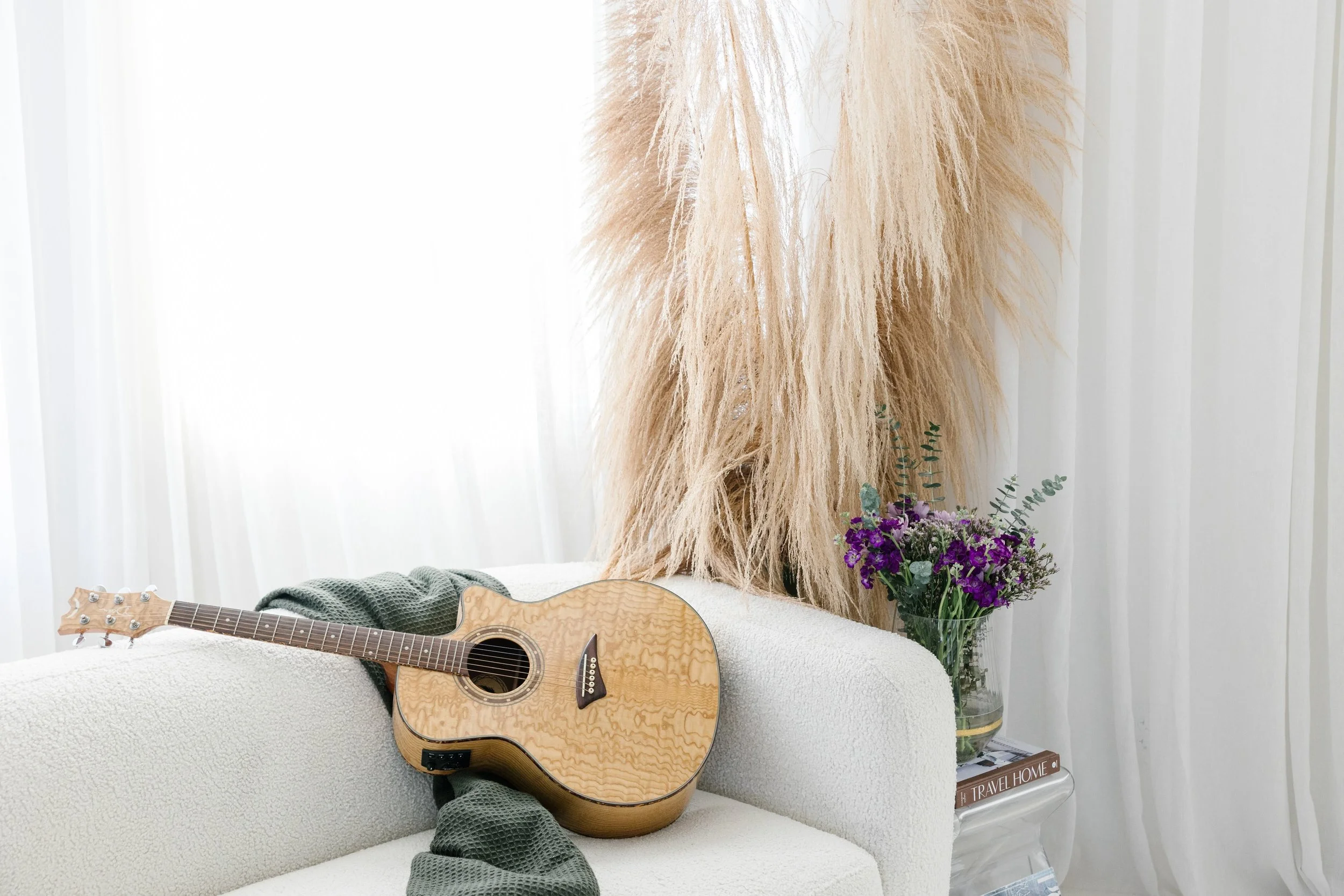 A cozy indoor scene with an acoustic guitar resting on a white couch, a green knitted blanket partially draped over it, beige pampas grass in a vase, and a bouquet of purple flowers on the table. White curtains in the background let in natural light.