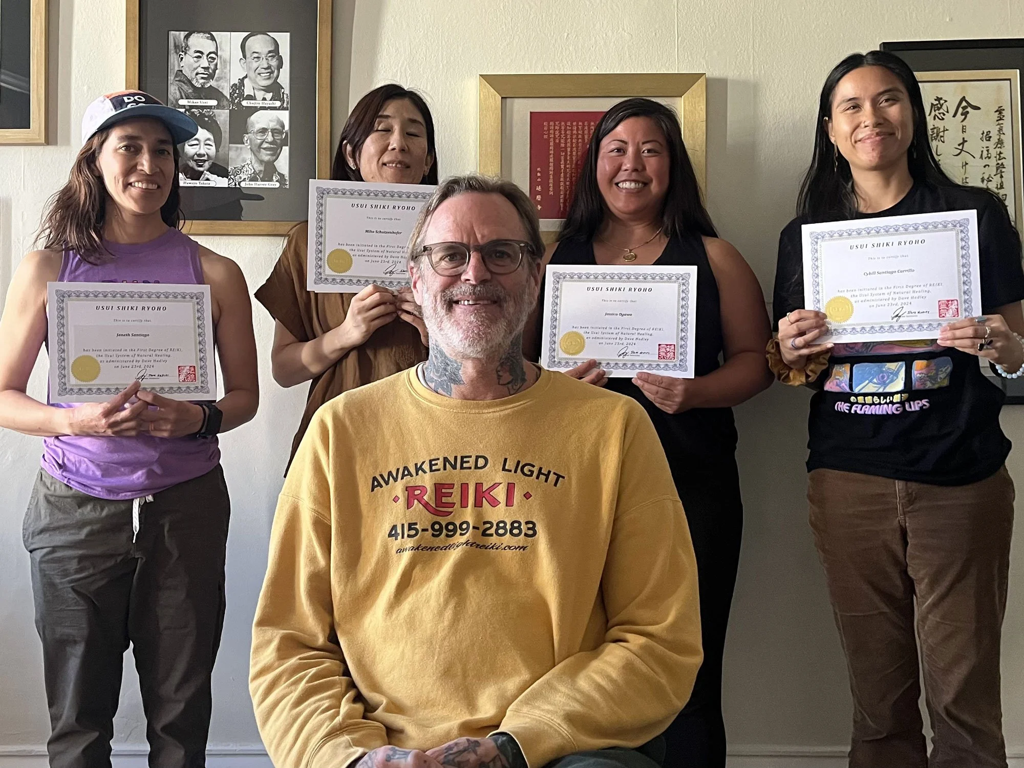 Group of five people, four women and one man, smiling and holding certificates, posing indoors against a wall with framed pictures and artwork.