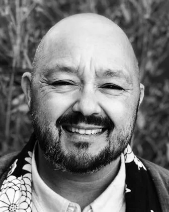 Smiling man with a bald head and beard, wearing a patterned shirt, outdoors with blurred trees in the background.