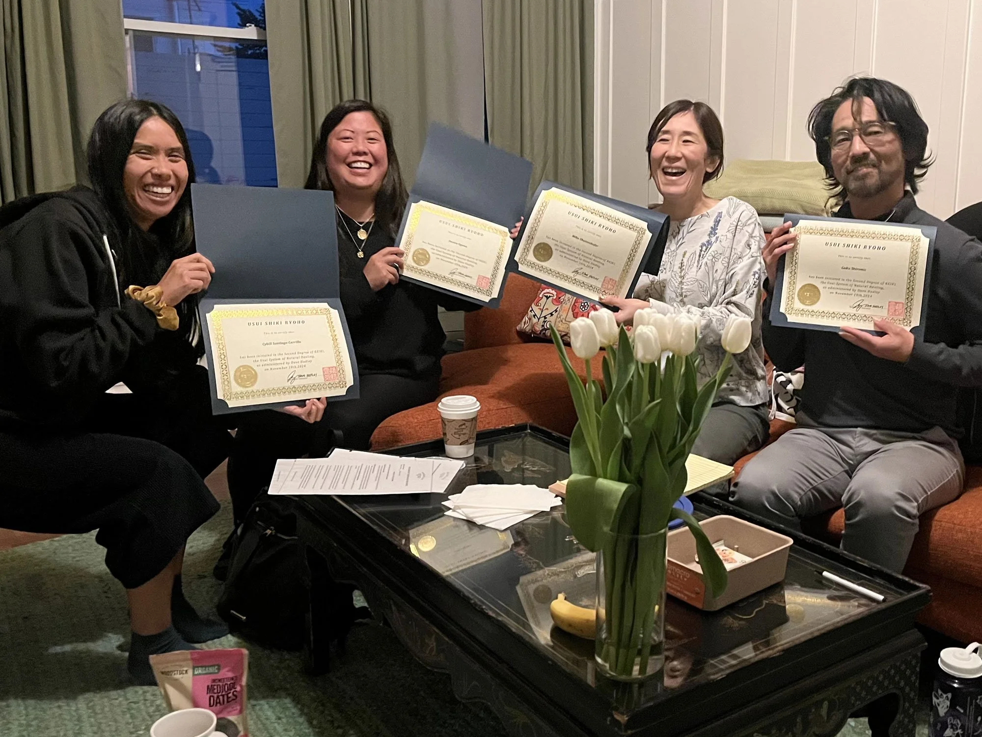 Five people sitting on a sofa in a living room, each holding a framed certificate and smiling. There are white tulips in a vase and a coffee table with various items in front of them.